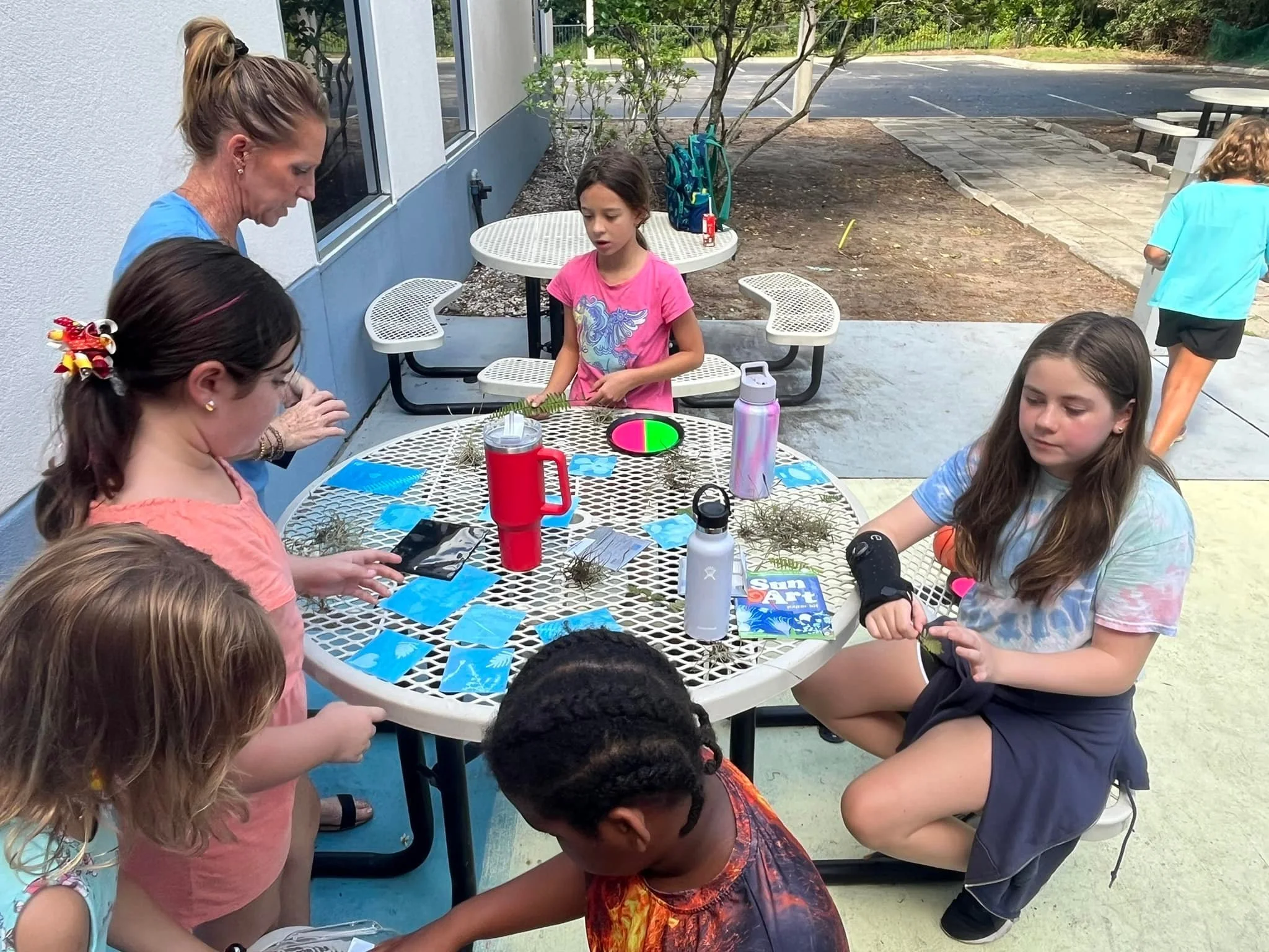 Children gathered around an outdoor table engaging in arts and crafts, with various art supplies and nature elements on the table.