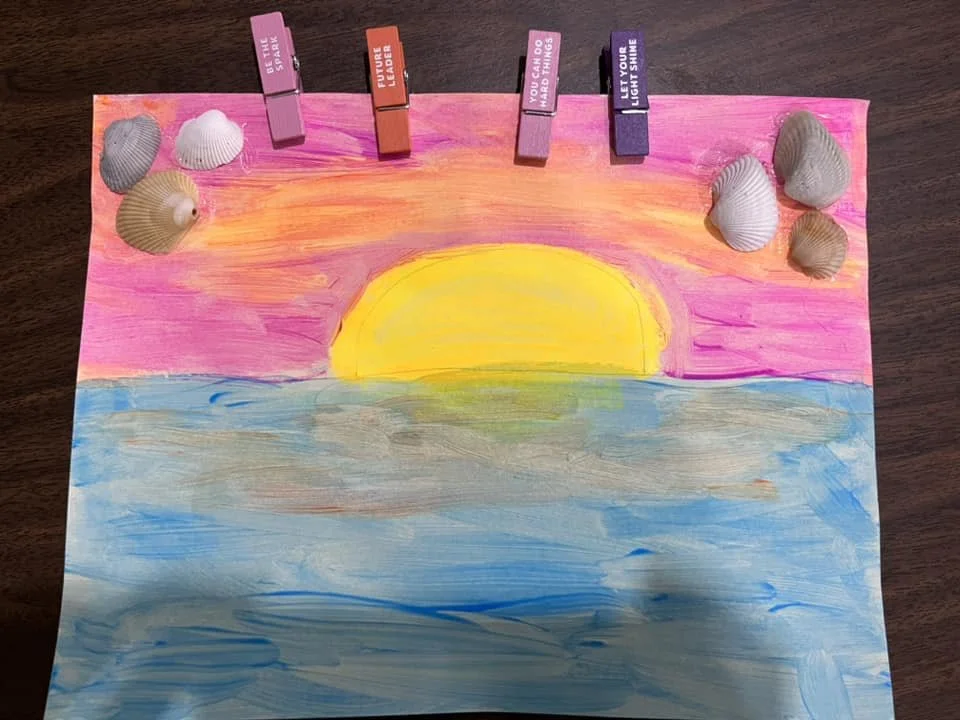 A hand-painted picture of a sunset over the ocean with pink and orange sky, yellow sun, and blue water. Seashells and clothespins with inspirational messages are attached to the top of the paper.