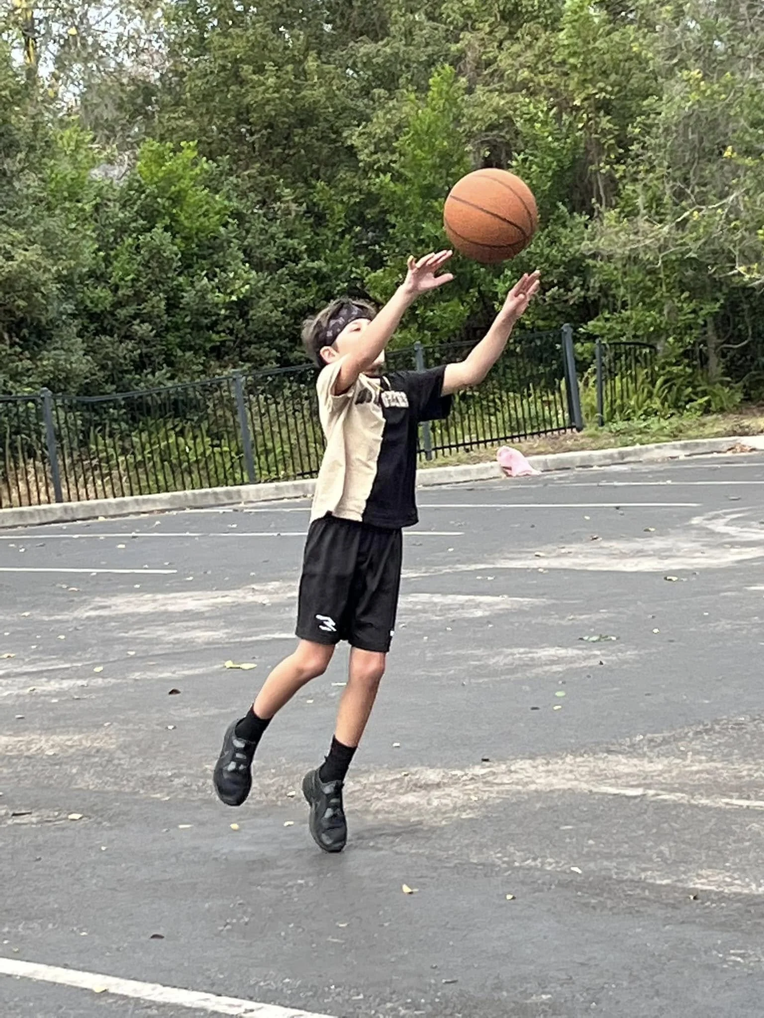 A boy playing basketball outdoors, jumping to shoot or catch the basketball, on a paved court with trees and bushes in the background.