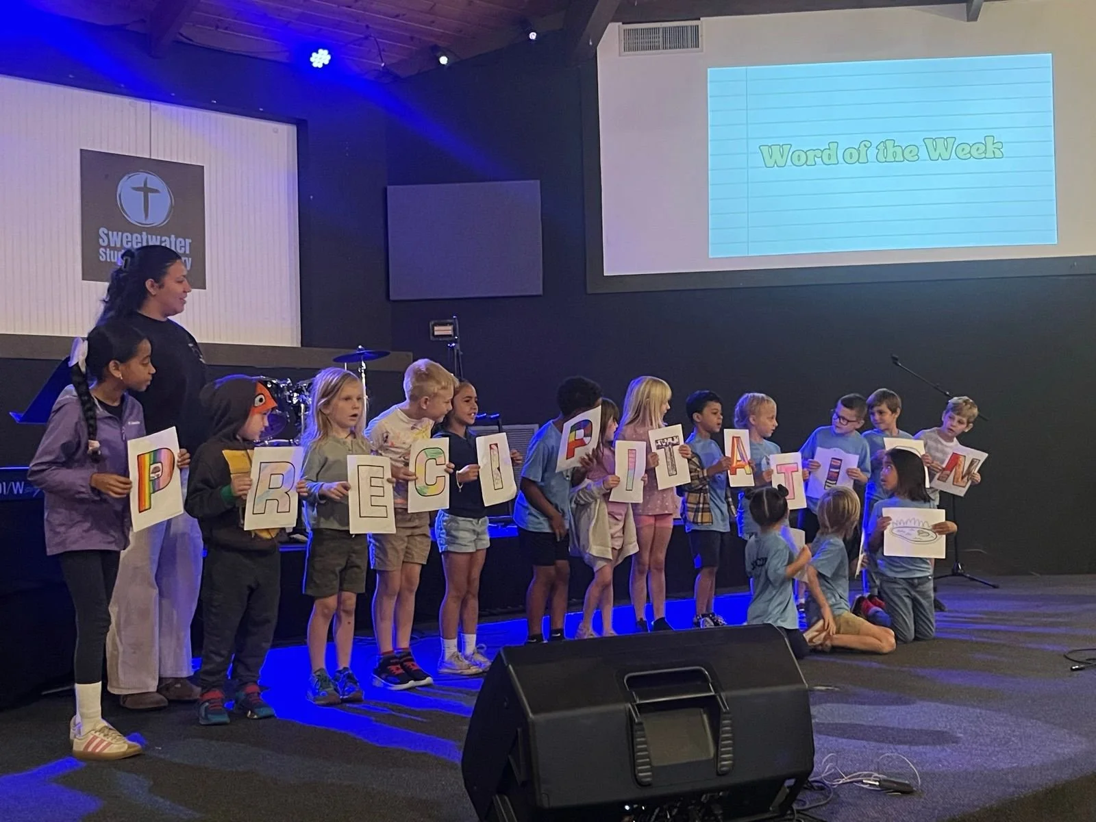 Children and a teacher on stage holding alphabet letter cards. A screen displays 'Word of the Week.' The backdrop shows 'Sweetwater' and a cross logo.