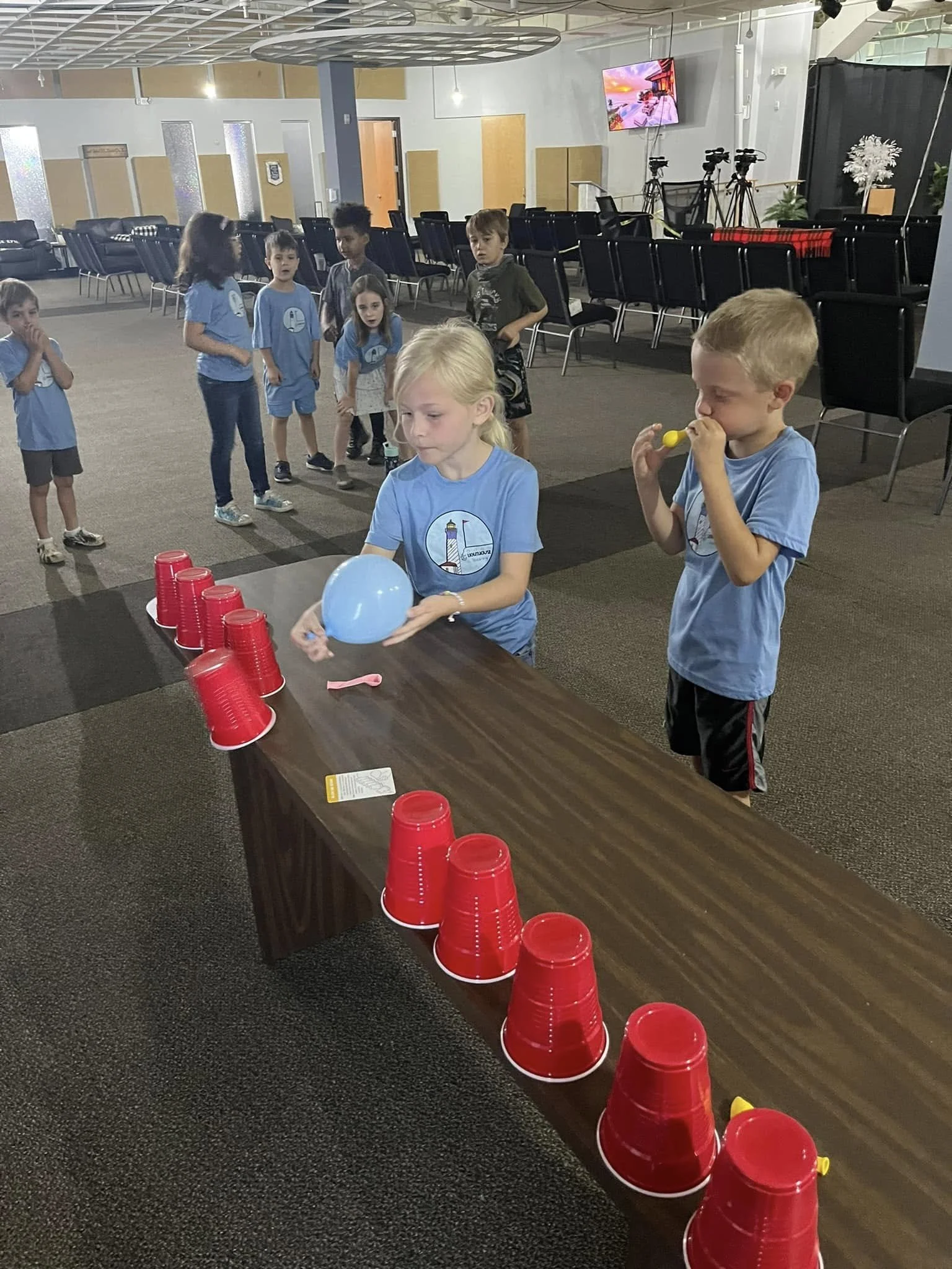 Children participating in a game where they stack red cups on a table; one girl is holding a balloon, and a boy is blowing a yellow whistle. Other children wait in line in the background inside a large indoor space with chairs, cameras, and a TV scre