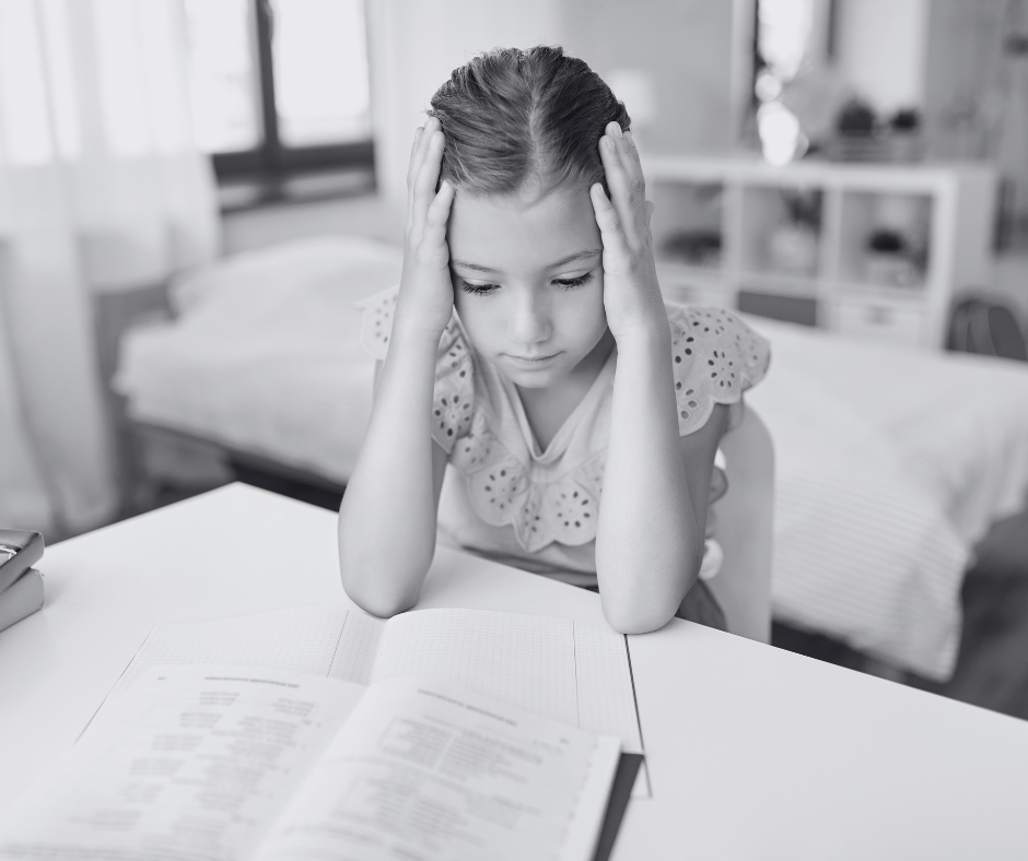 A young girl sitting at a desk with her head in her hands, looking down at an open book.
