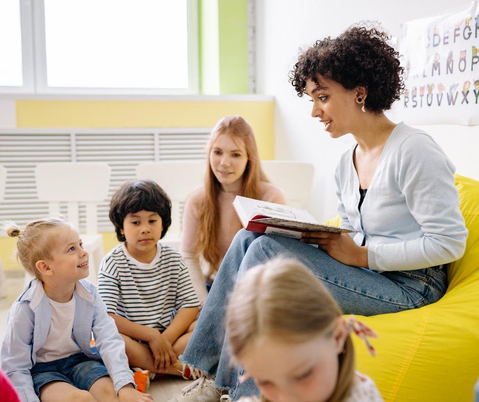 An adult woman reads a book to four young children sitting on the floor in a classroom.