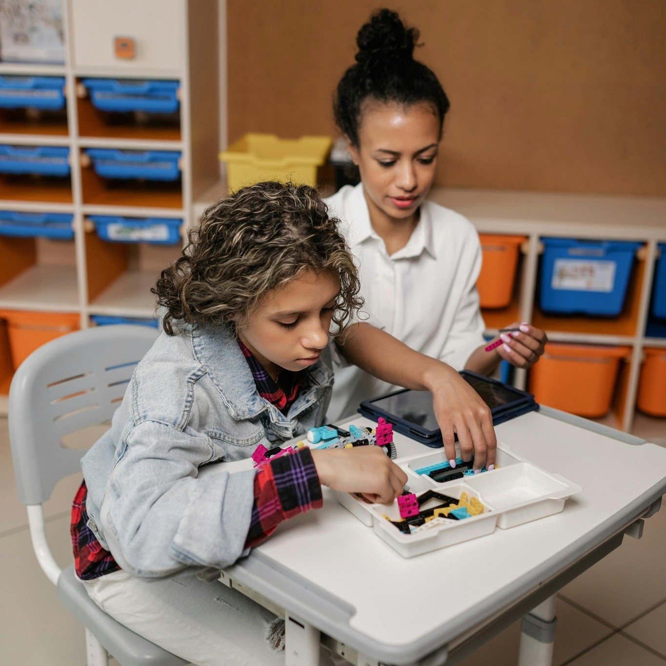 Two girls working with LEGO robotics pieces at a school table in a classroom with cubby storage in the background.