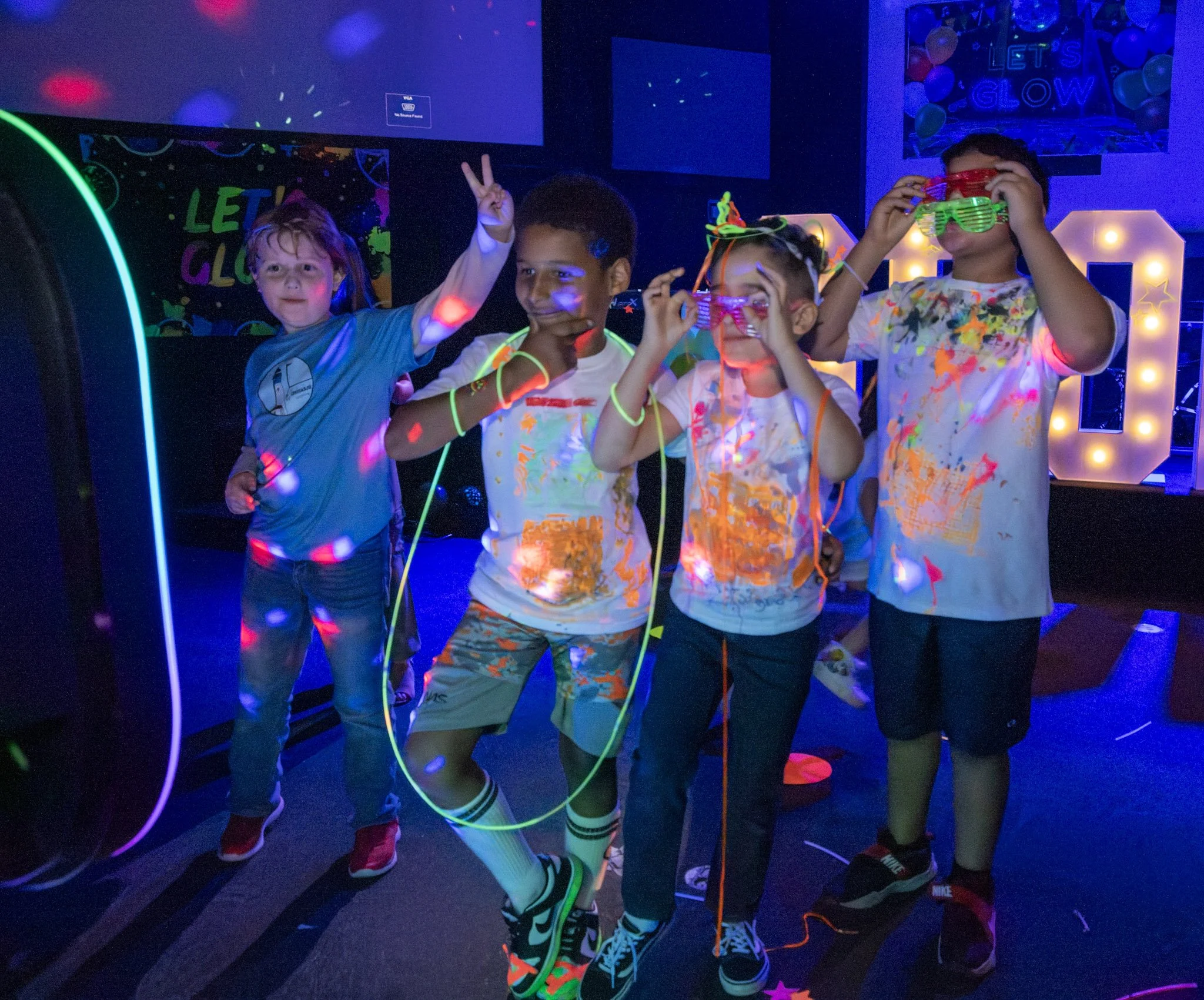 Children in a dark room with blacklight, wearing glow-in-the-dark clothes and glasses, celebrating at a party with neon decorations.