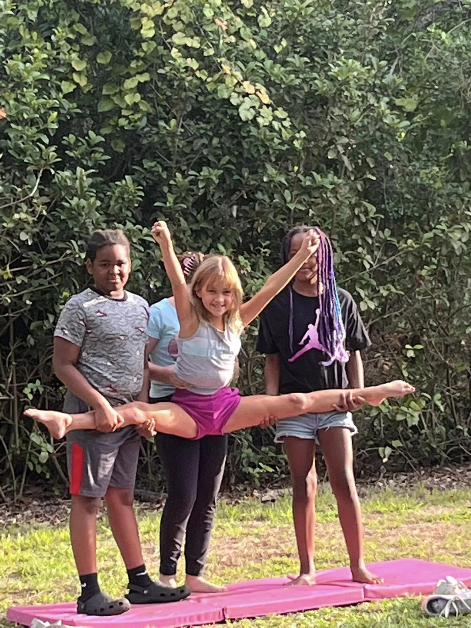 Four children outdoors on a pink mat, with green trees in the background. One girl with blonde hair and a light-colored tank top is performing a split while smiling. She is supported by a girl with long braided hair, a black t-shirt with a pink graph
