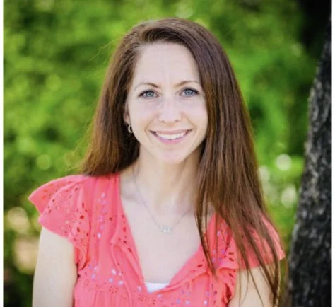 A young woman with long brown hair and blue eyes smiling outdoors, wearing a pink top with lace details, with green foliage in the background.