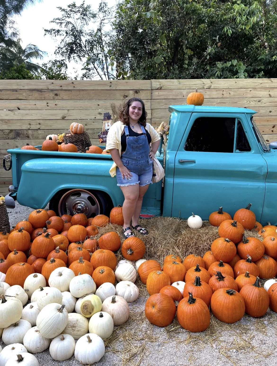 A woman standing next to an old blue pickup truck surrounded by orange and white pumpkins, with some pumpkins in the truck bed. The scene is set outdoors with a wooden fence and greenery in the background, suggesting a fall or harvest theme.