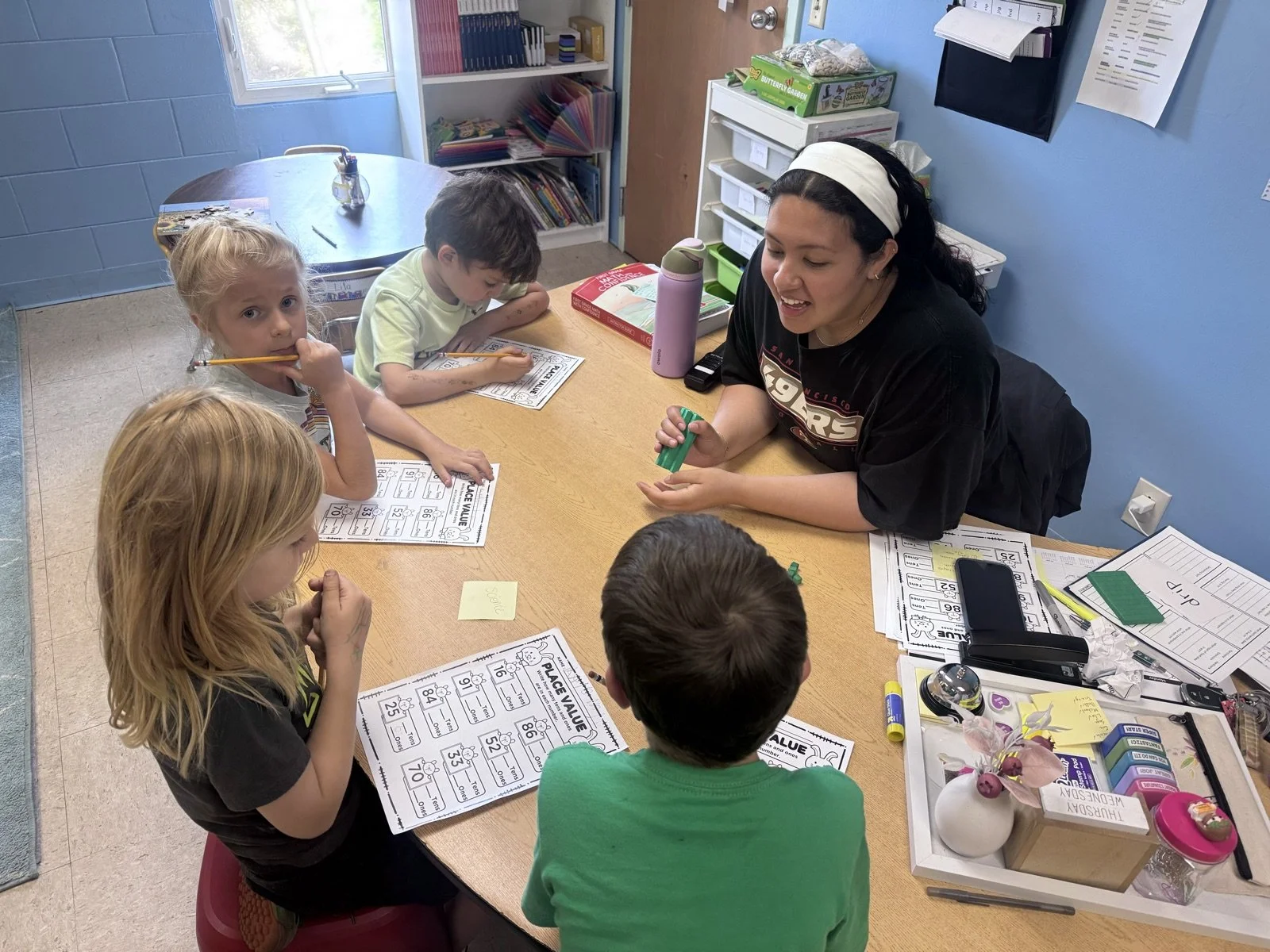 In this classroom scene, a teacher is engaging with young students at a table, distributing a small green object while the children work on worksheets related to place value.
