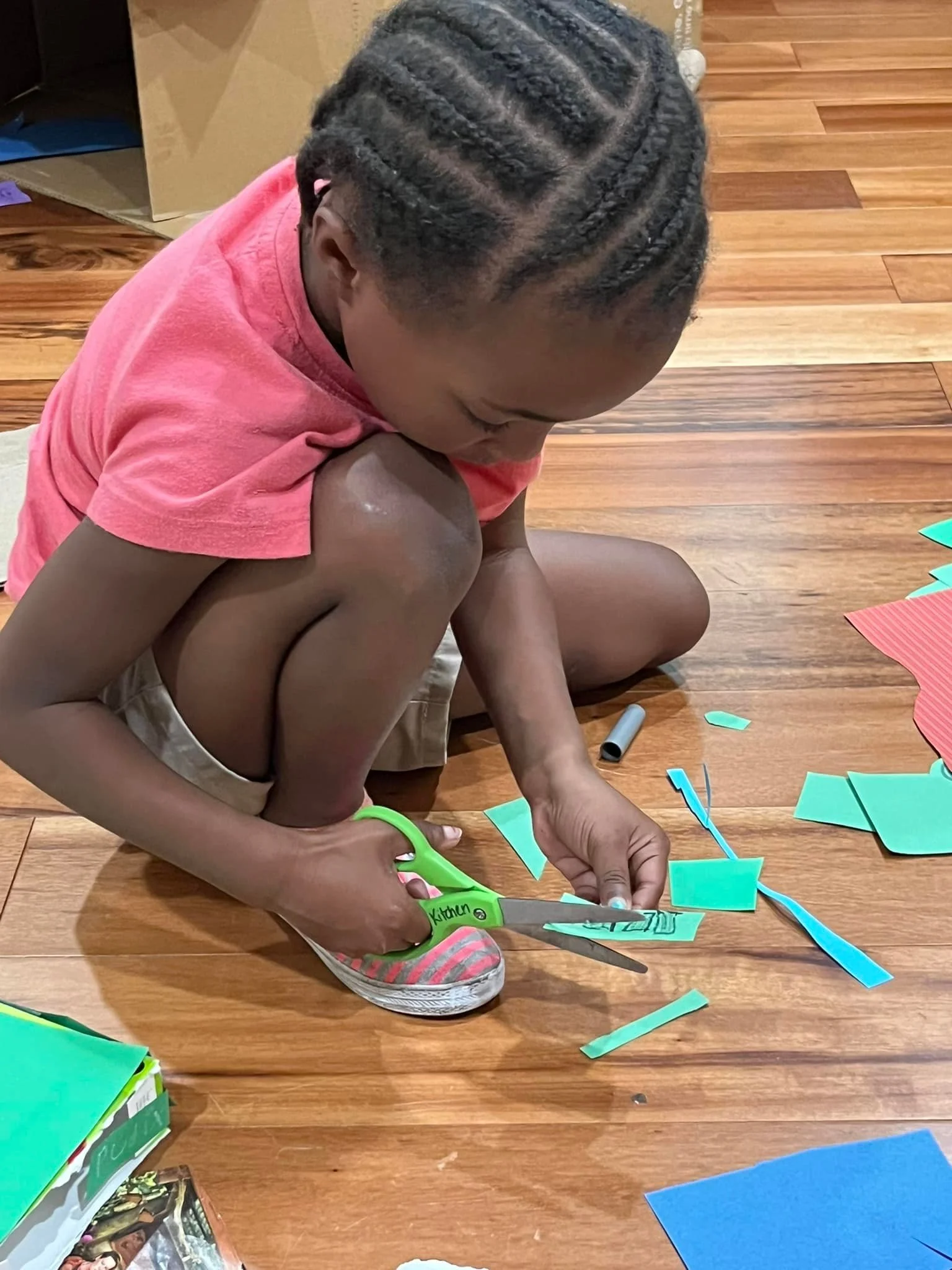 A young boy with braided hair, wearing a red t-shirt, is sitting on a wooden floor, cutting pieces of colored paper with green scissors for a craft project.