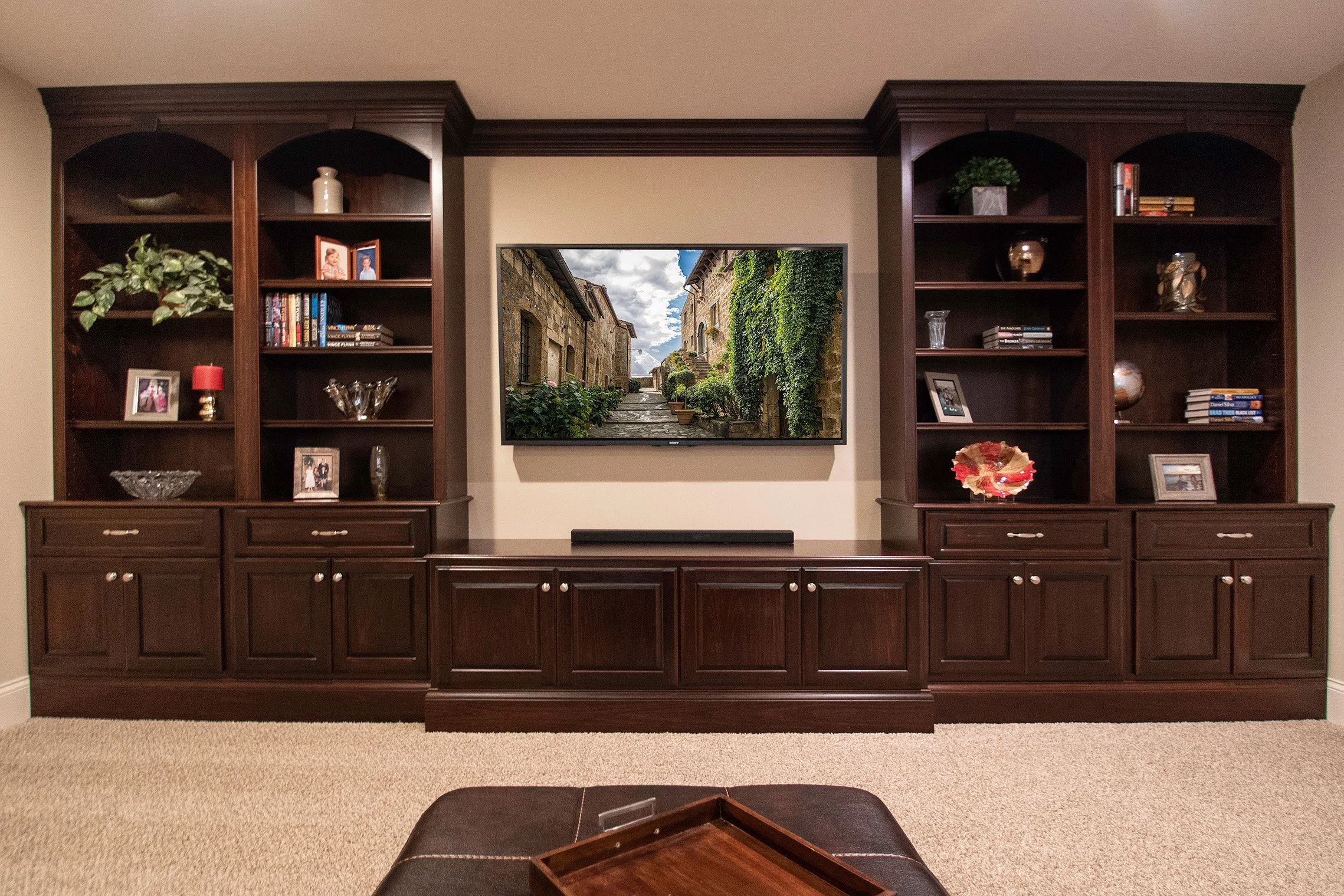 Living room with dark wood built-in shelves, a wall-mounted TV displaying an outdoor scene, and a beige carpeted floor.