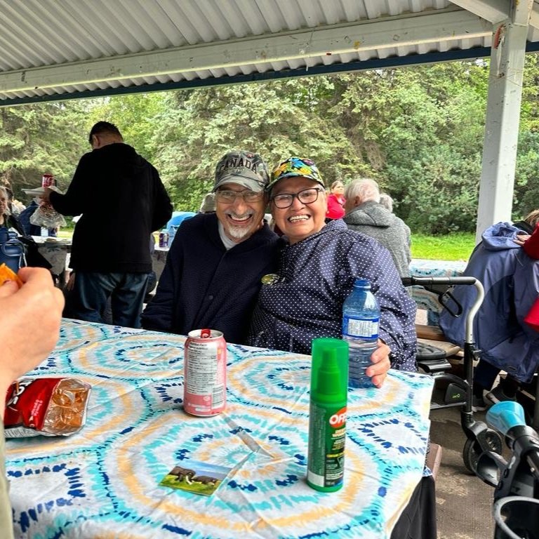  A man and a women sit at a picnic table smiling. They have their arms around each other and both are wearing glasses and ball caps.  