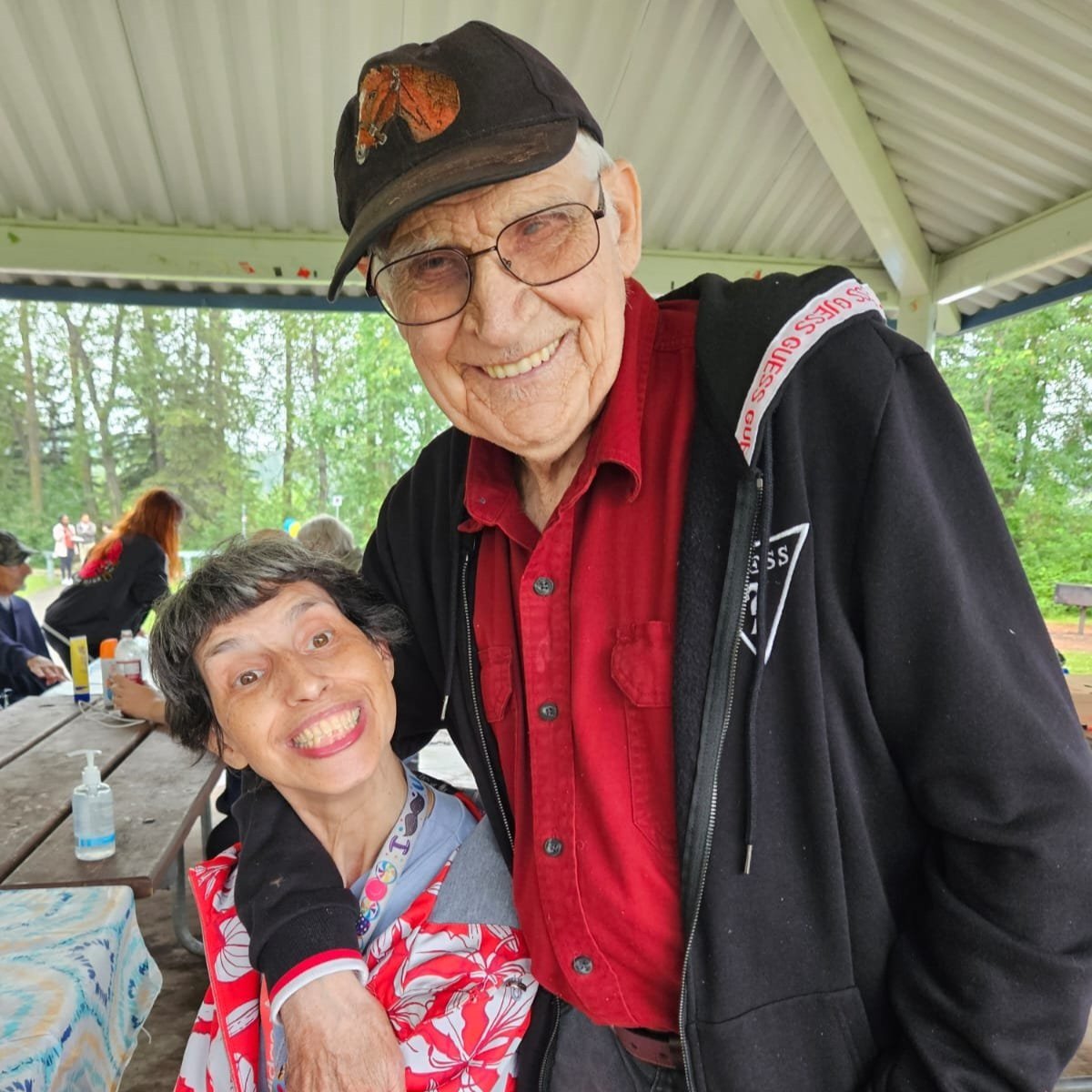  A man and women with their arms around each other are smiling. The women is seated at a picnic table. The man is wearing a ball cap and glasses.  