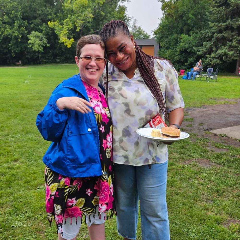  Two women smile with their arms around each other. One women holds a plate of food.  