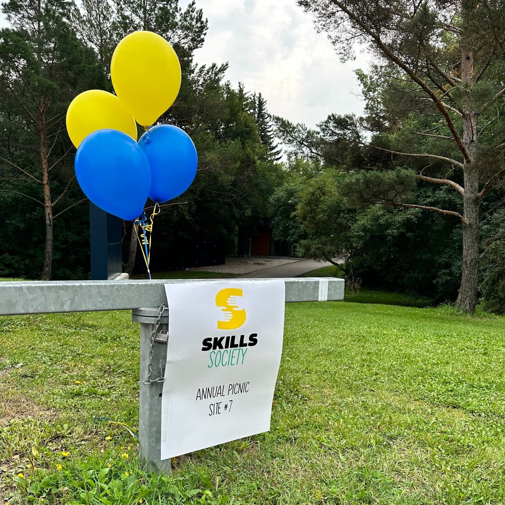  A sign for the Skills Society picnic is attached to a parking barrier. Four balloons, two yellow and two blue are floating above it.  