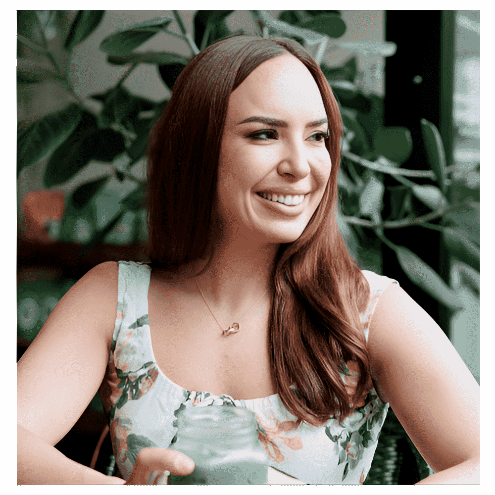 A smiling woman with long auburn hair sitting indoors surrounded by large green plants.