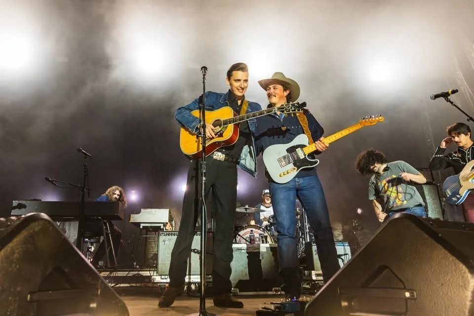 Two musicians, a woman with blonde hair in a denim jacket and a man wearing a cowboy hat and a dark shirt, perform on stage with guitars, surrounded by band members with musical instruments. They are smiling and enjoying the moment.