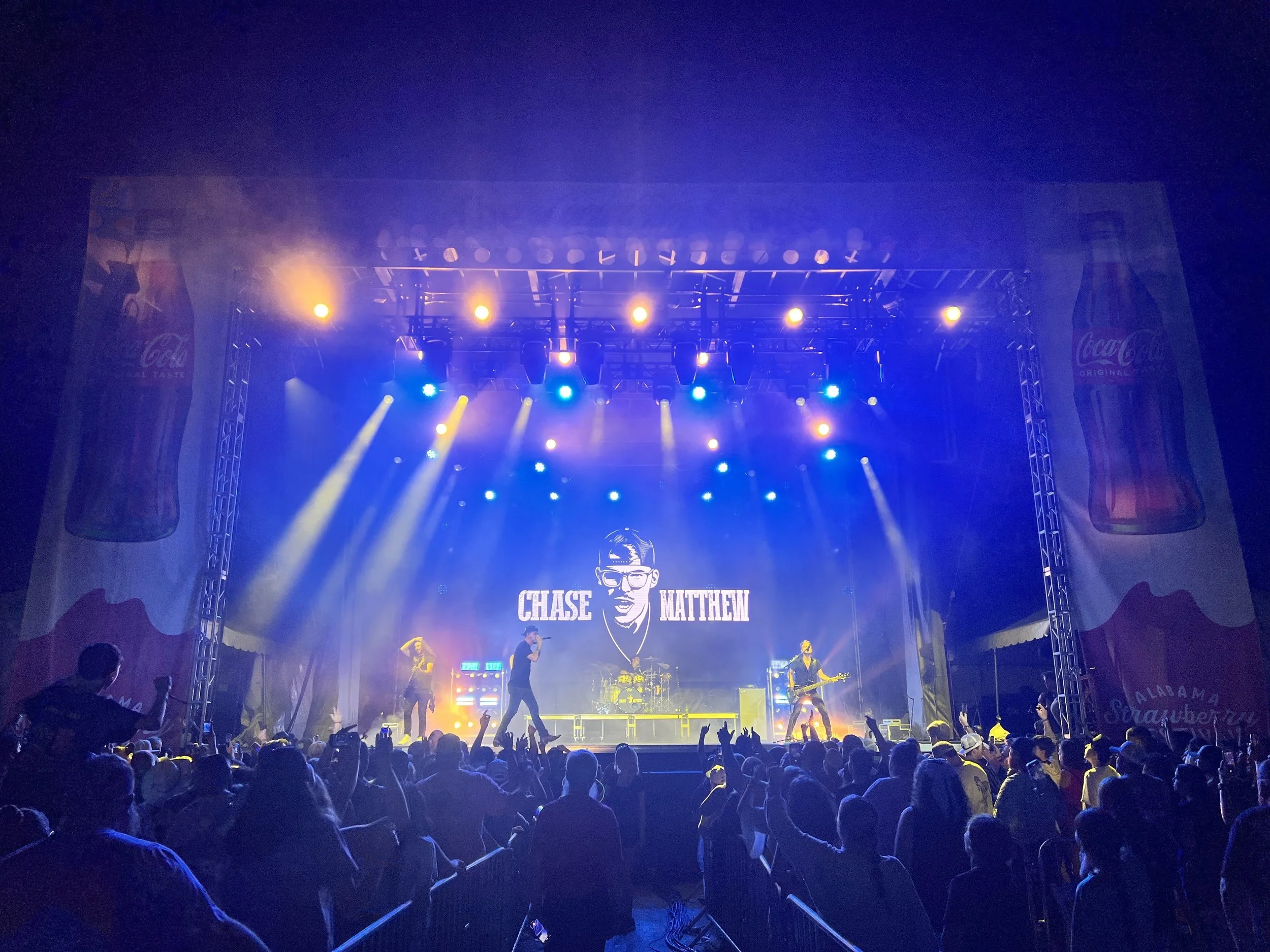 Concert stage with band performing, large crowd with some raising hands, blue and yellow stage lights, screens displaying 'Chase Matthew', Coca-Cola banners on sides.