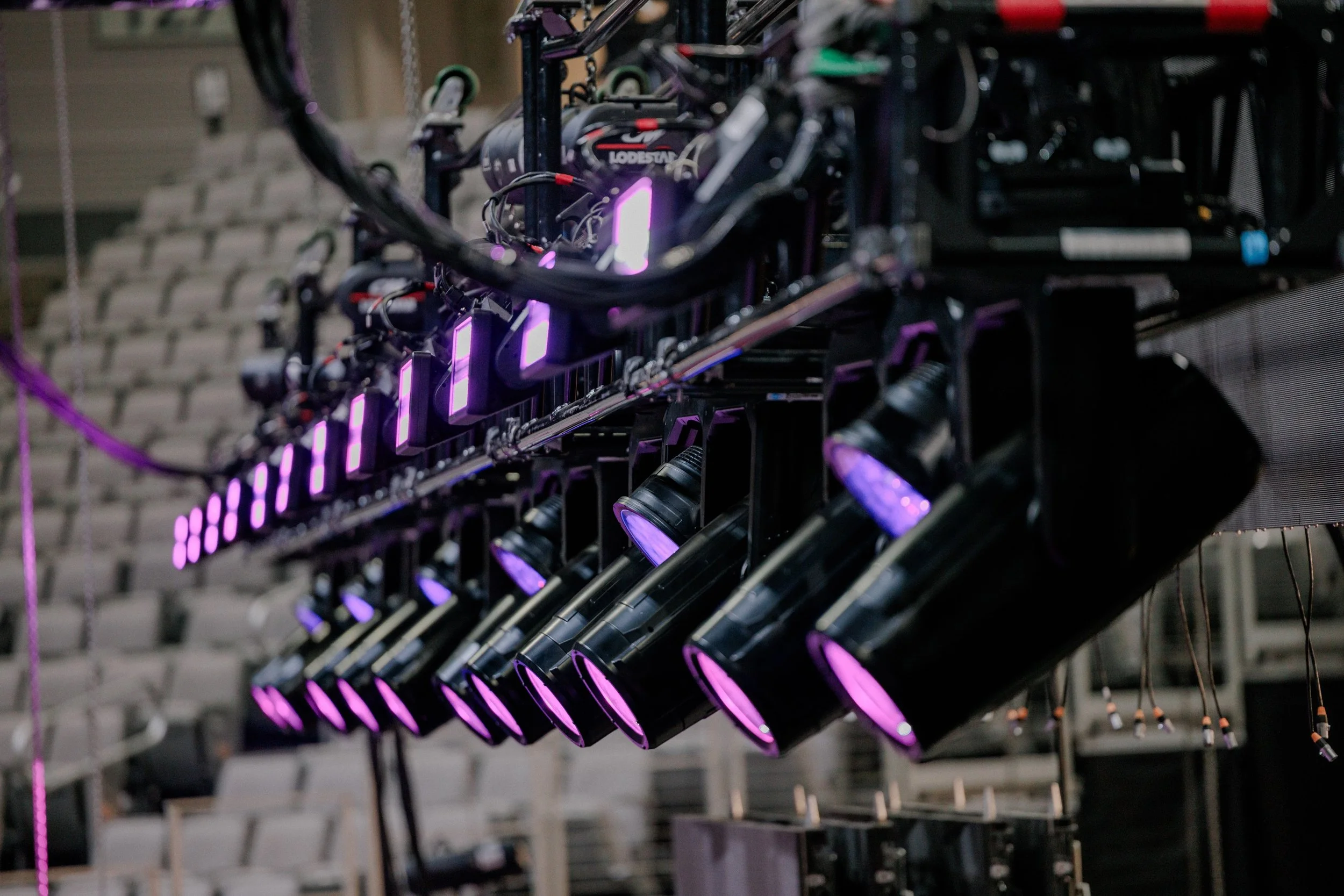 A row of professional stage lighting equipment with purple lights, mounted on a rig in a theater or concert venue.