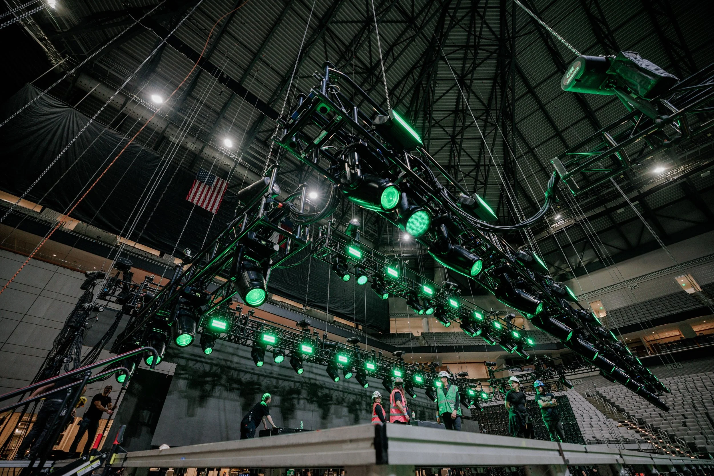 Large stage lighting rig with green lights, hung above a group of workers setting up the stage in an indoor arena with empty seating and American flag.
