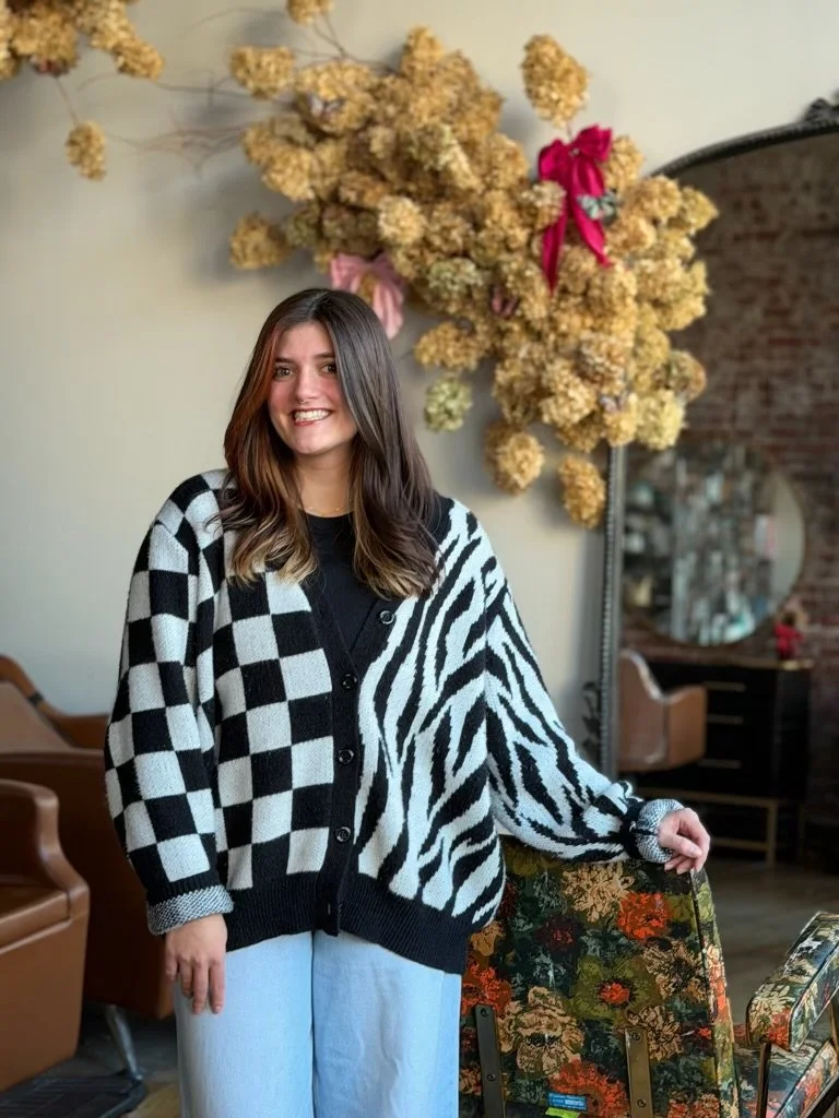A young woman smiling and standing indoors near a floral-patterned chair, decorated with a large arrangement of dried yellow flowers and pink ribbons, with a mirror and brick wall in the background.