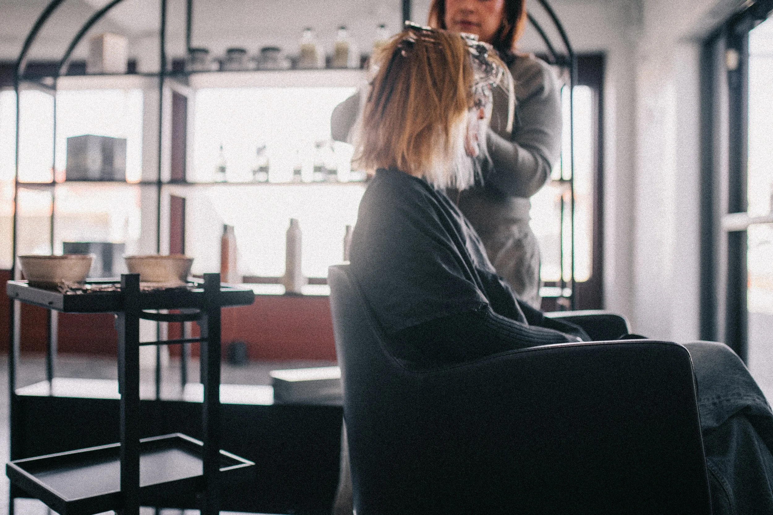 A hairdresser applying hair dye to a client's hair in a salon with large windows in the background.