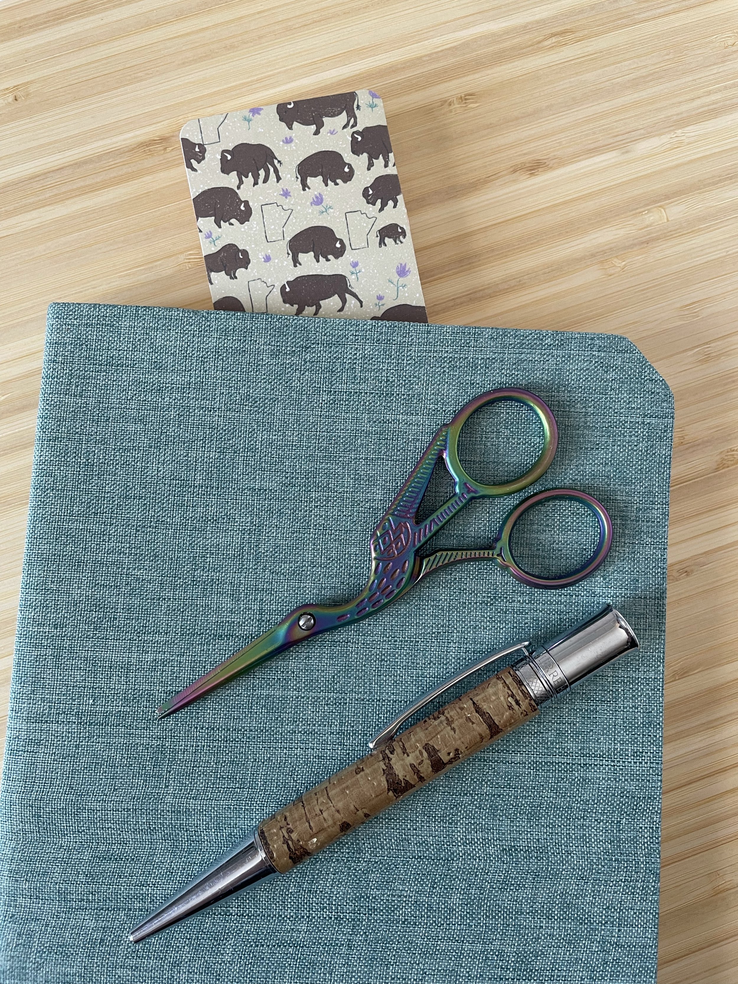 A bookmark featuring bison pops out the top of a book. Ontop of the book lies a pen and a pair of embroidery scissors.