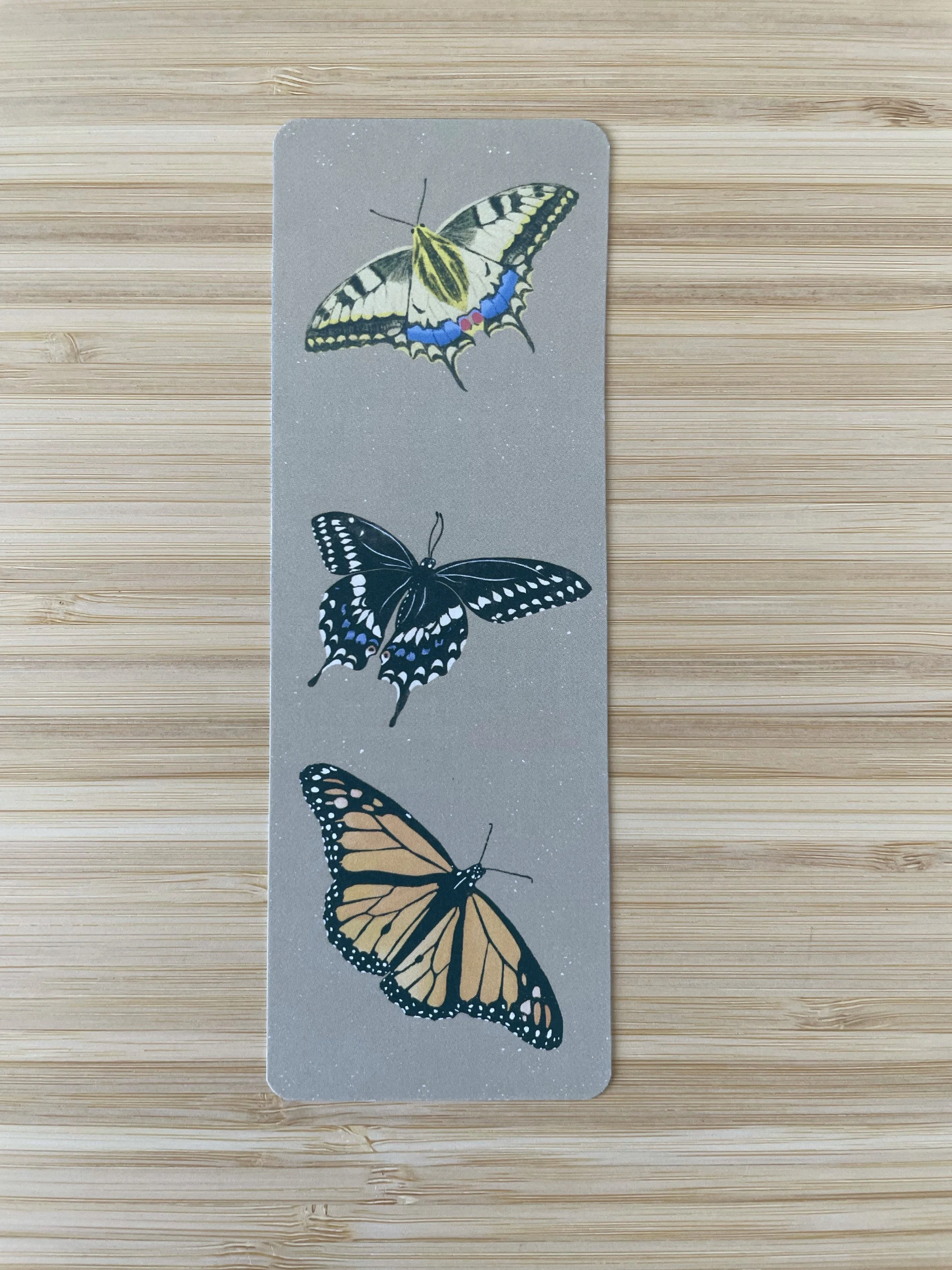 A tan bookmark featuring a Canadian Tiger Swallowtail, Eastern Swallowtail and Monarch Butterfly, sits on a wooden surface.