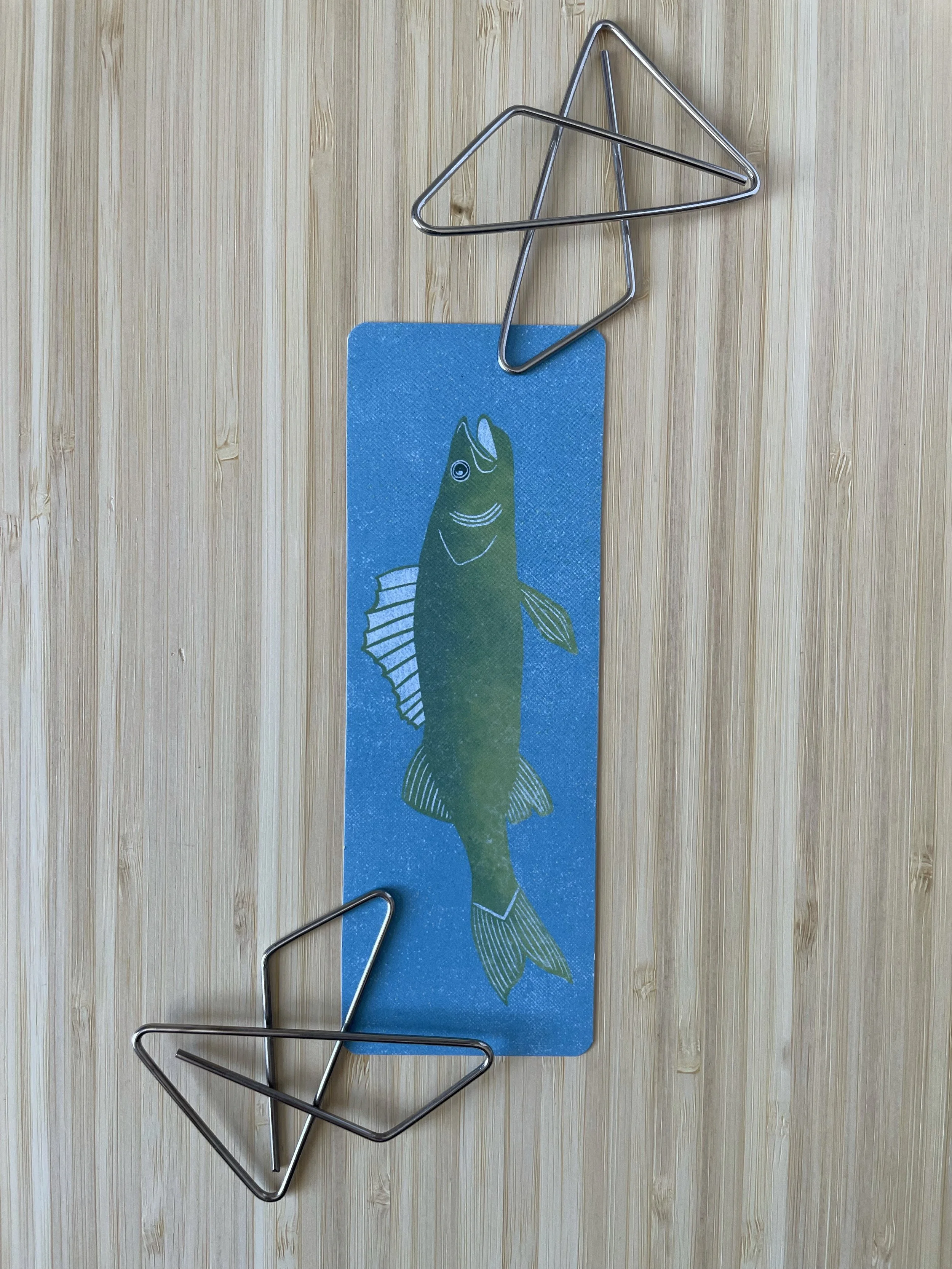 A bookmark featuring a pickerel sits on a wooden surface with two large paperclips.