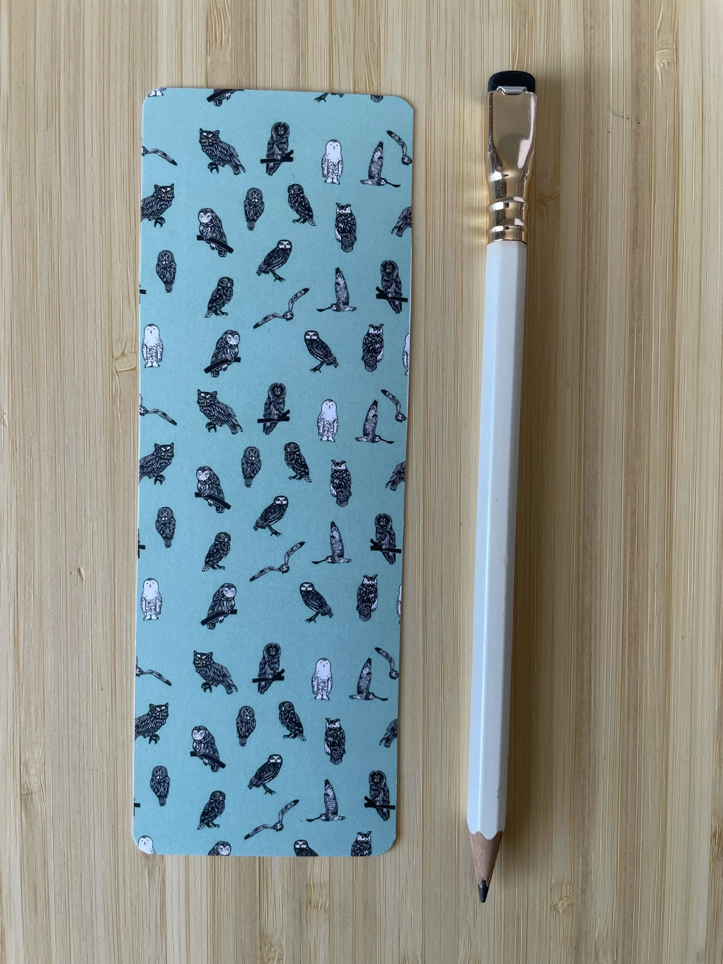 A light teal bookmark dotted with mini owls sits beside a white pencil on a wooden background.