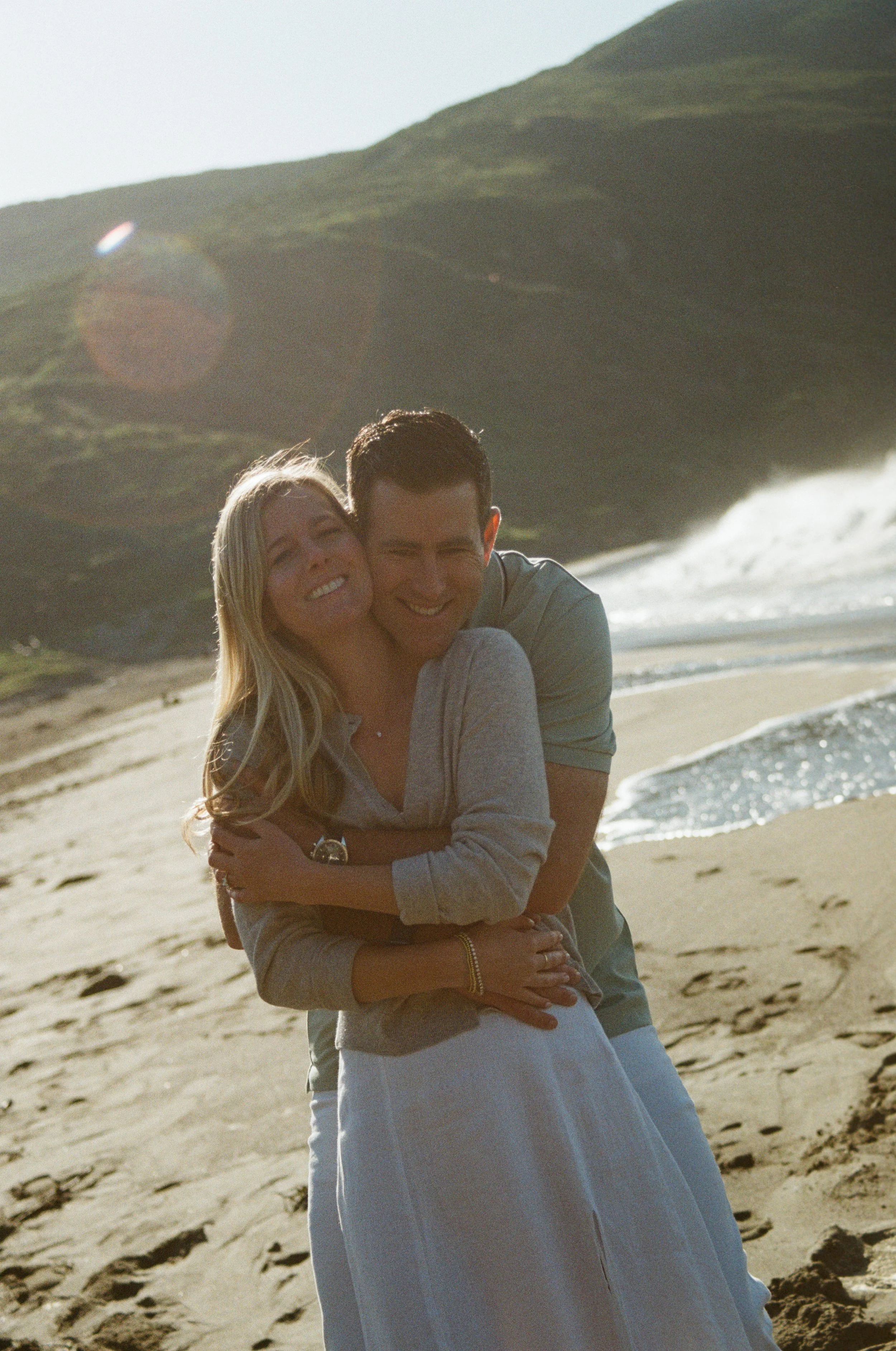 A couple embracing and smiling on a sandy beach with ocean waves and green hills in the background, during late afternoon sunlight.