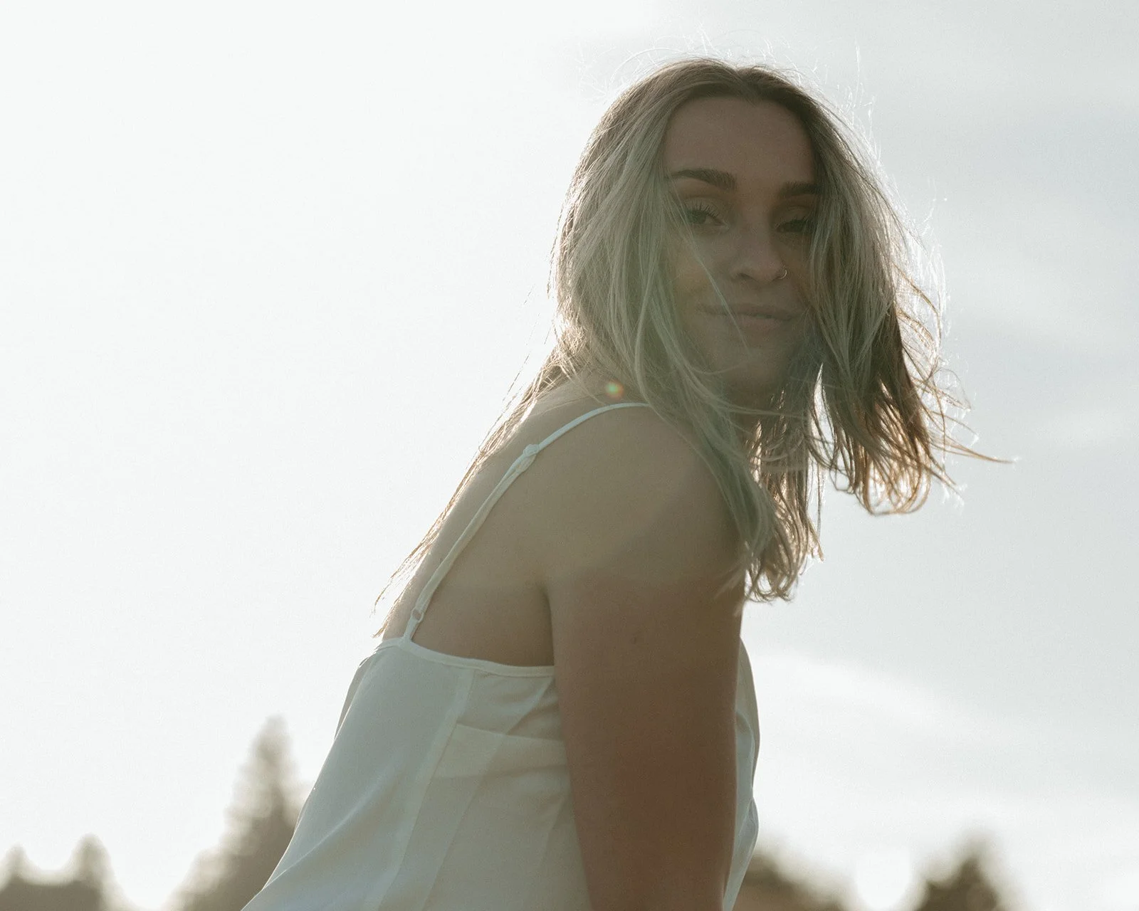 A young woman with wavy blonde hair wearing a white top, smiling softly outdoors with a bright sky in the background.