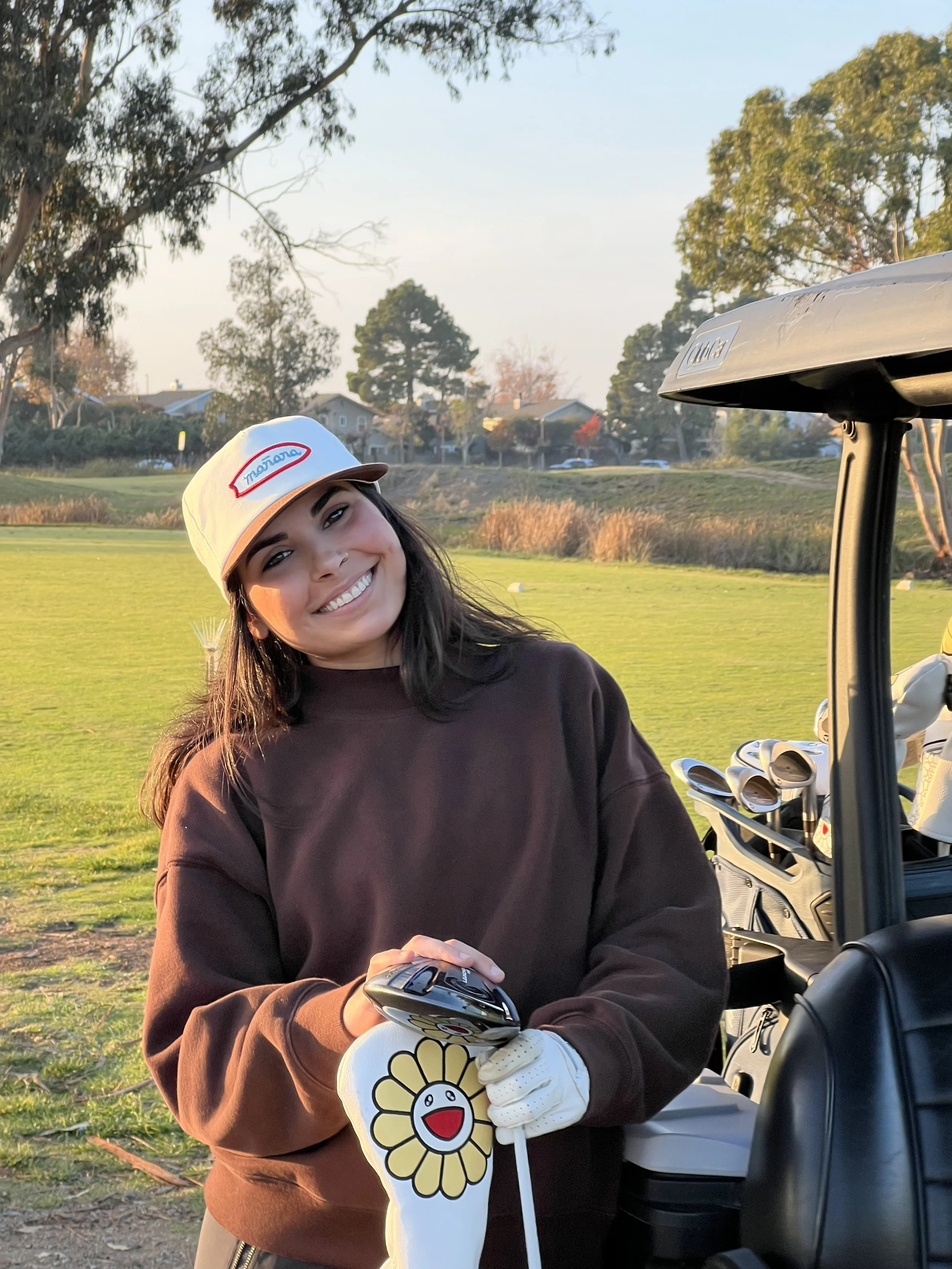A young woman smiling on a golf course, holding a golf club and wearing a white cap with a red and blue logo, and a brown sweatshirt. Golf cart with clubs is nearby. Trees and houses are in the background during sunset.