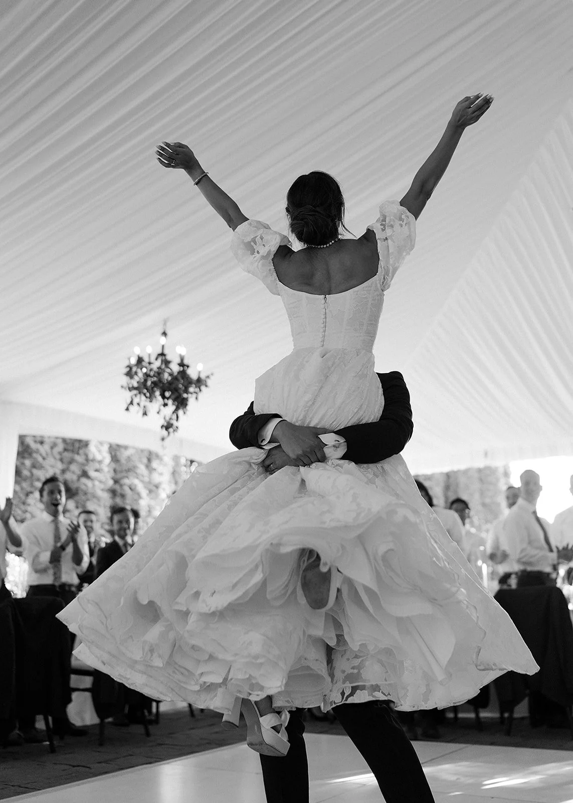 A bride in a wedding dress is being lifted by her groom during a dance at a wedding reception. The bride has her arms raised and is spinning, with guests clapping and watching in the background.