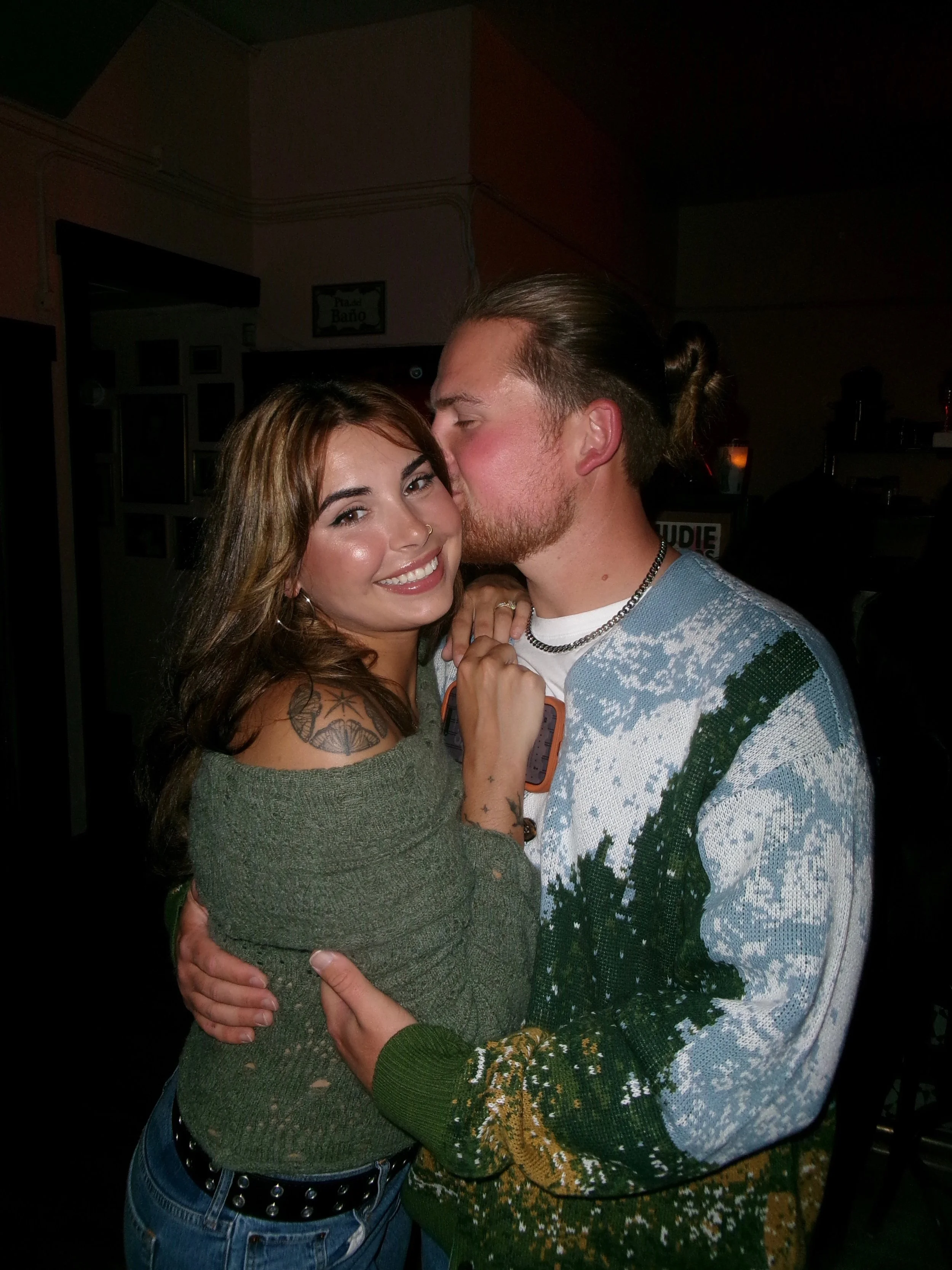 A young woman with tattoos on her arm smiling and hugging a young man with long hair tied back, who is kissing her cheek in a dimly lit indoor setting.