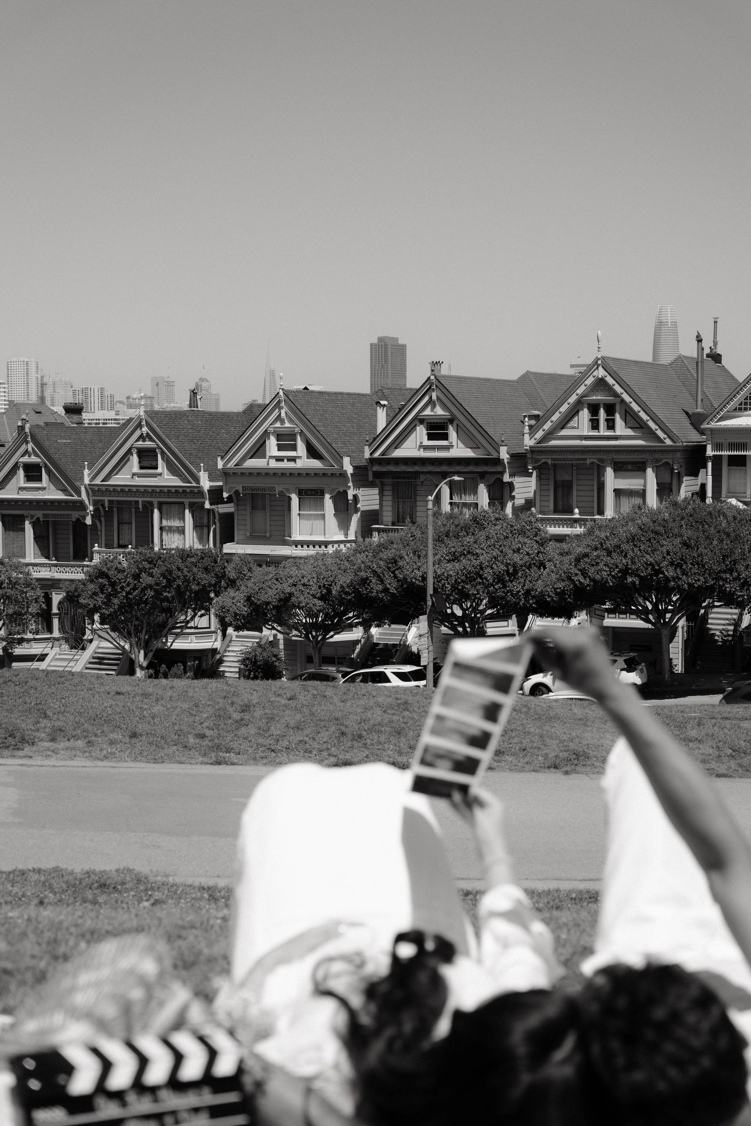 A person lying on the grass holding a movie clapperboard with a city skyline in the background, featuring Victorian-style houses in the foreground.