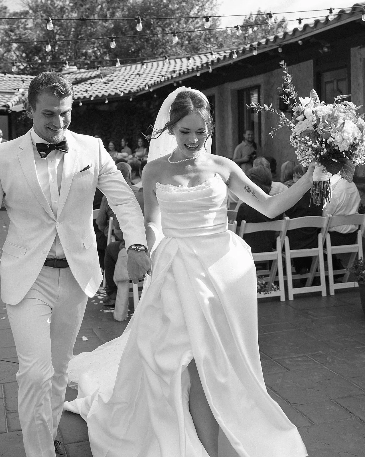 A joyful bride and groom walking hand in hand at their outdoor wedding reception. The bride is holding a bouquet, smiling, and wearing a strapless wedding dress, while the groom is dressed in a white suit with a bow tie. Guests are seated in the background under string lights.