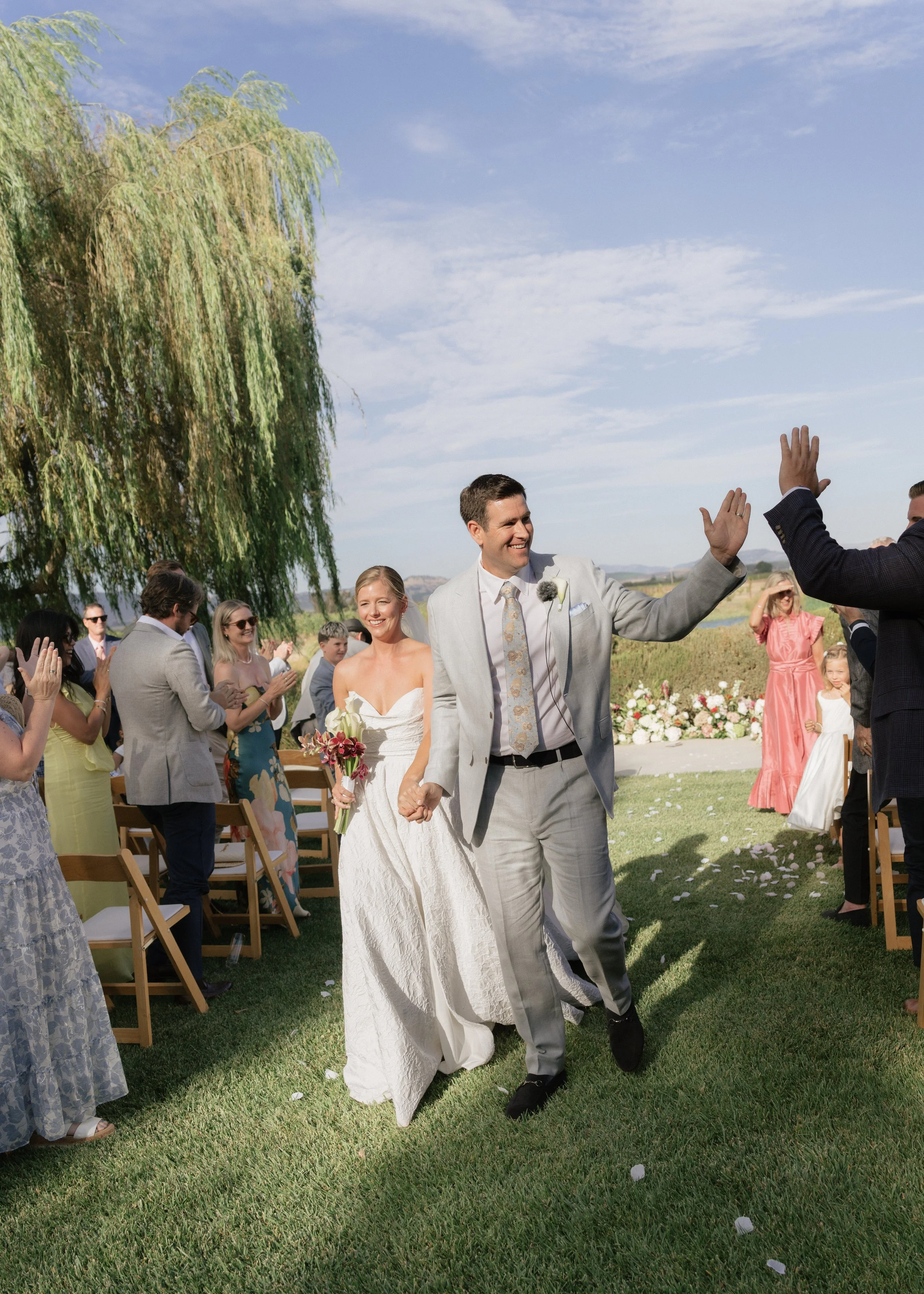A newlywed couple holding hands and smiling as they walk down a grassy outdoor aisle during their wedding ceremony. The bride is in a white dress holding a bouquet of flowers, and guests are clapping and smiling on either side of the aisle.