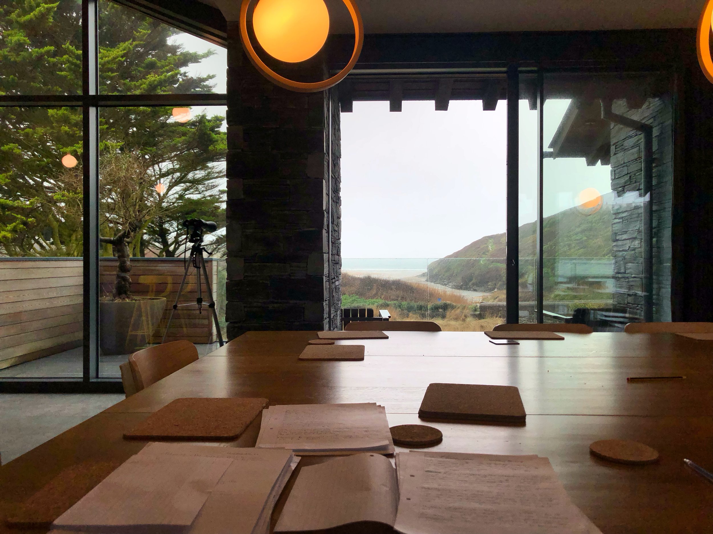 Interior view of a room with a wooden table and papers, overlooking a scenic outdoor view with a large window. Outside, there are trees and a tripod visible, with a glimpse of a hilly landscape and the sea in the distance. Pendant lights hang from th