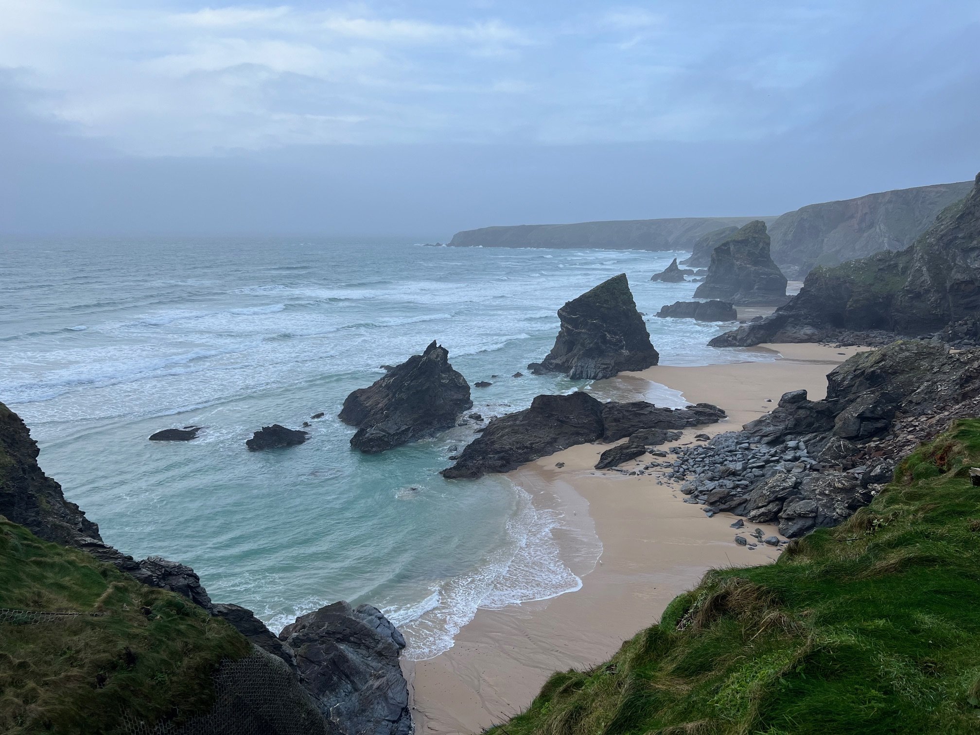Rocky coastline with sandy beach and large cliffs at Bedruthan Steps in Cornwall, UK, under a cloudy sky.