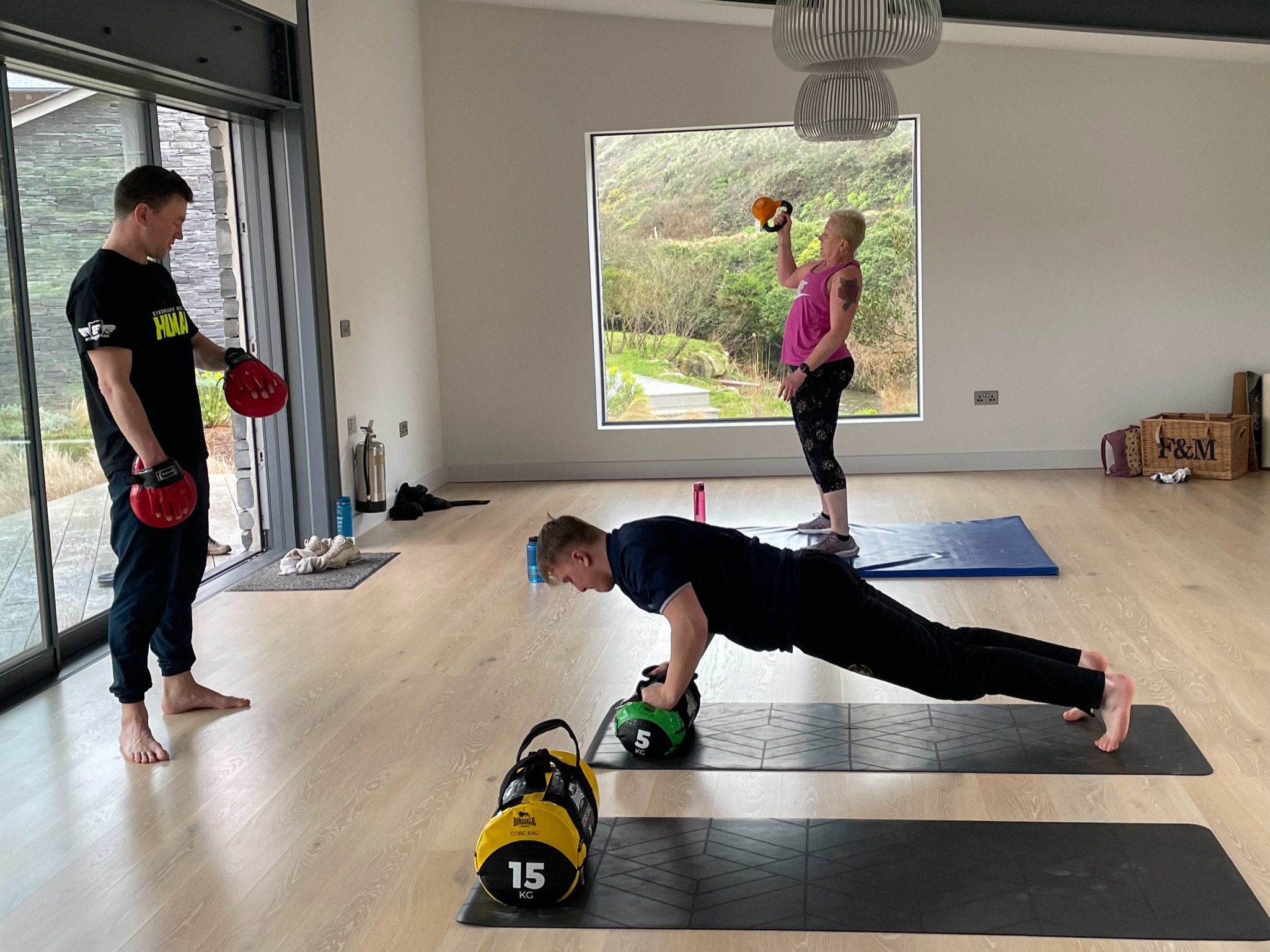 A home gym session with three people. One person performs a plank using a kettlebell, another holds a kettlebell overhead while standing on a mat, and the third person stands nearby with training pads. The room features large windows overlooking gree