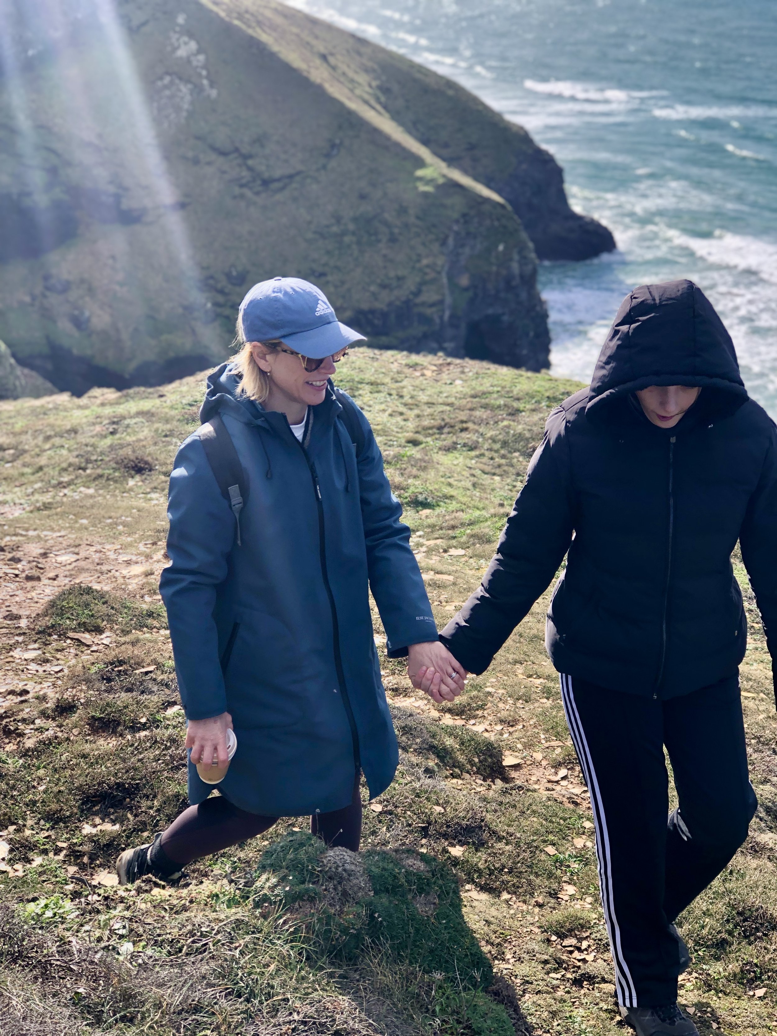 Two people walking on a grassy cliffside path near the ocean, holding hands. One is wearing a blue coat and cap, the other a black hooded jacket.
