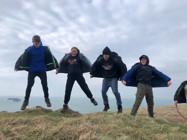 Four people jumping in the air on a grassy hill with a cloudy sky and ocean in the background.