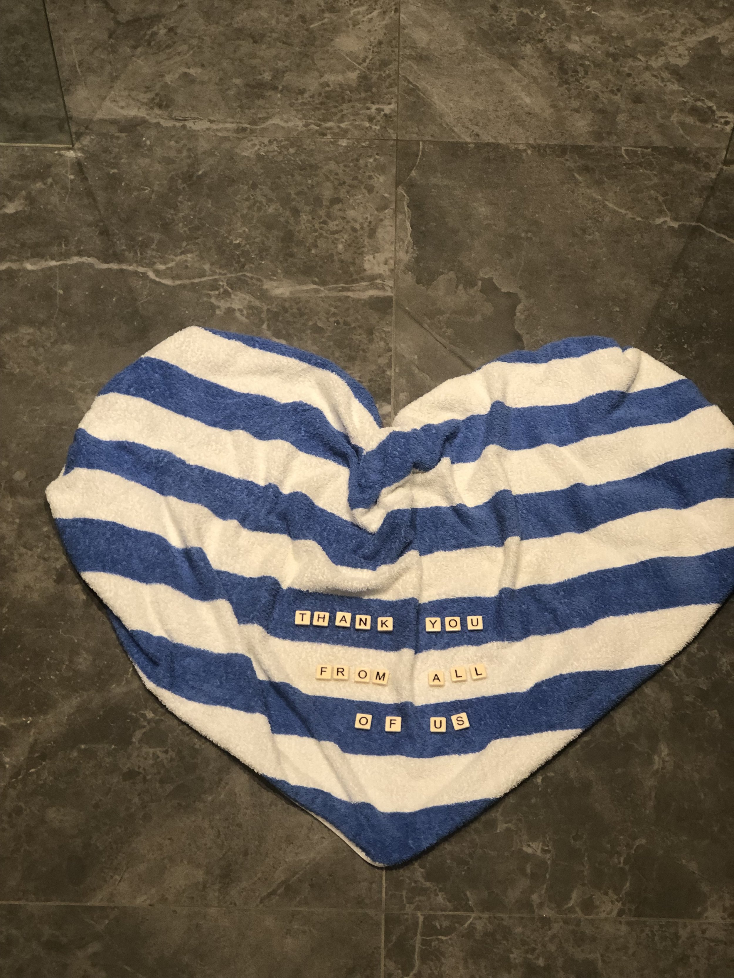 Blue and white striped towel folded into a heart shape with tiles spelling "Thank you from all of us" on it, placed on a gray tile floor.
