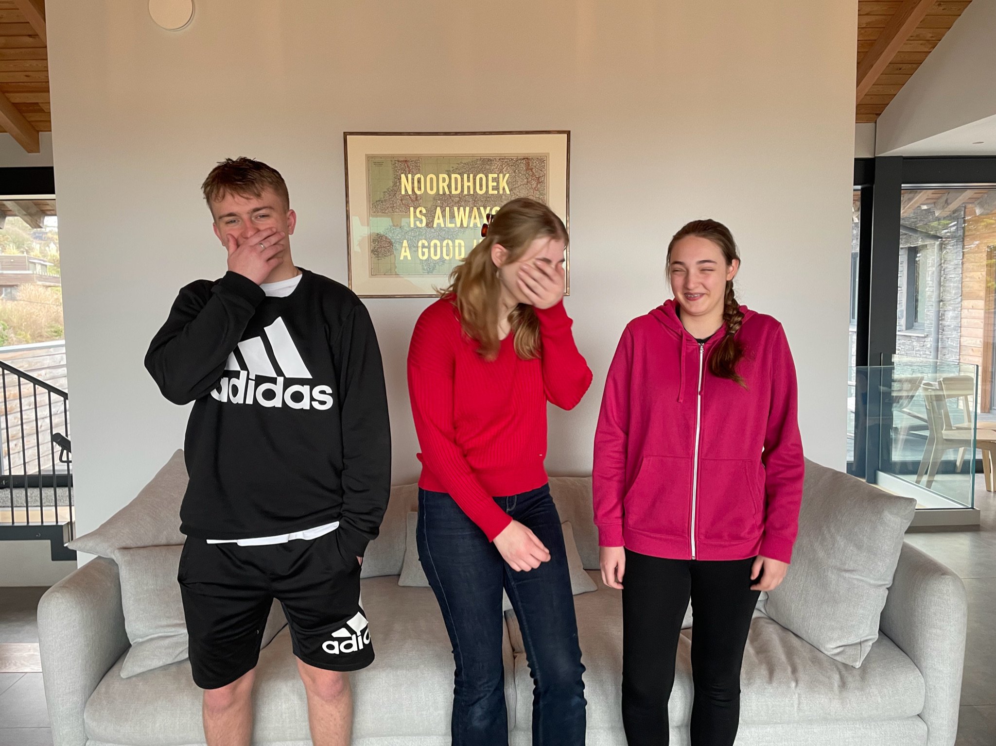 Three people standing and laughing indoors in front of a "Noordhoek is always a good idea" sign, with casual clothing.