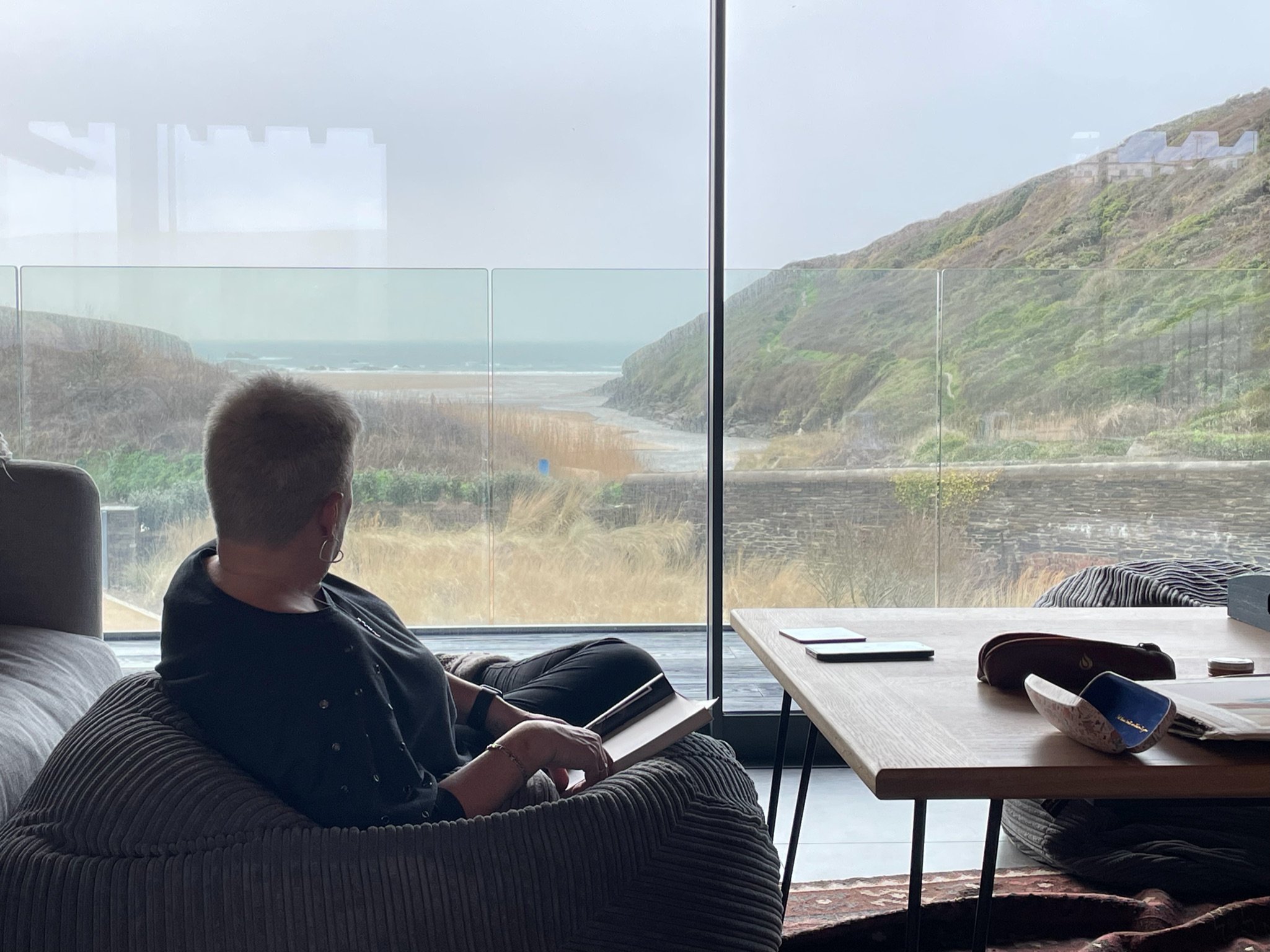 Person sitting indoors on a sofa, looking out at a coastal view through large glass windows, with hills and the ocean in the background.