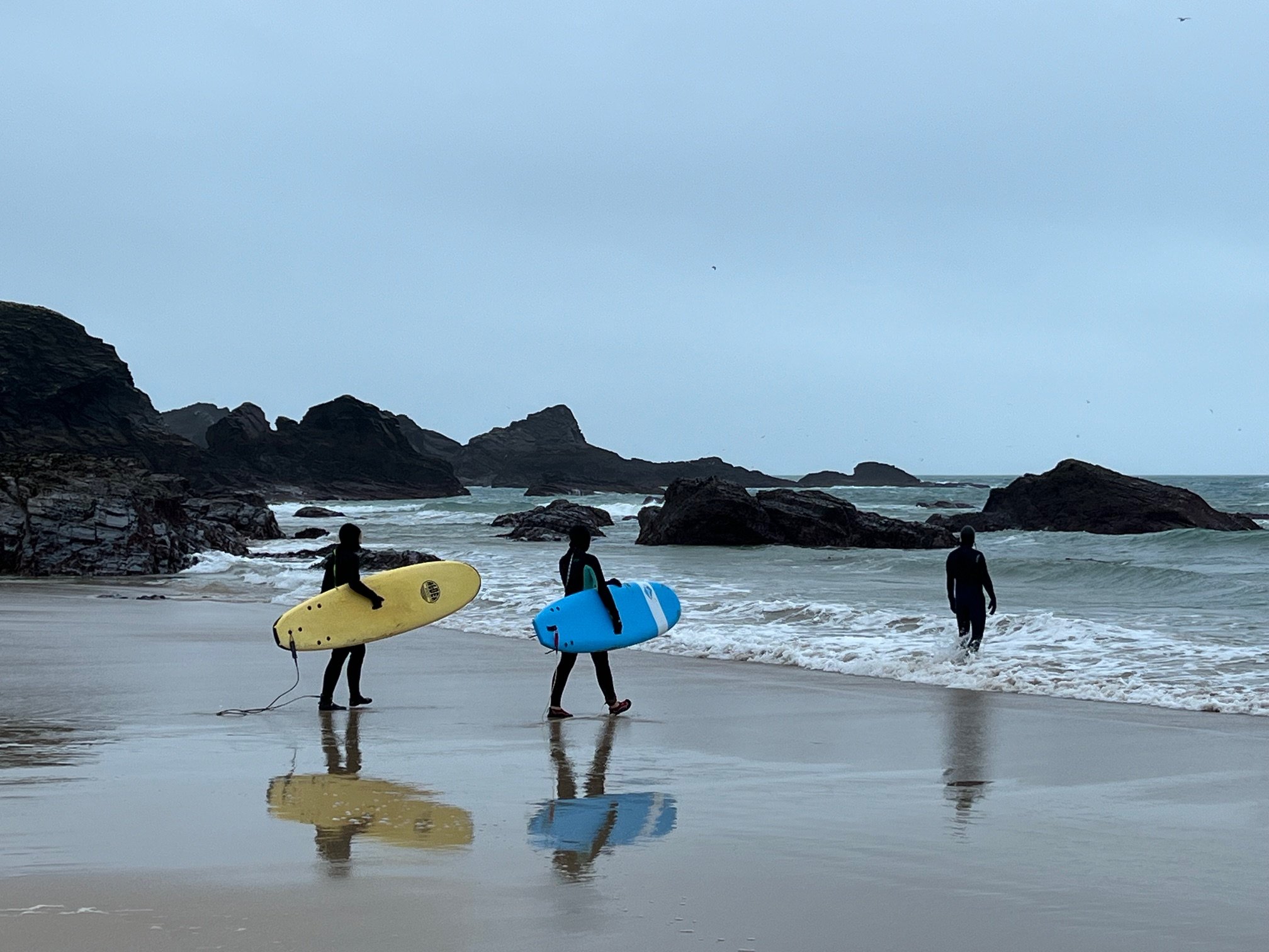Three people with surfboards walking on a rocky beach.