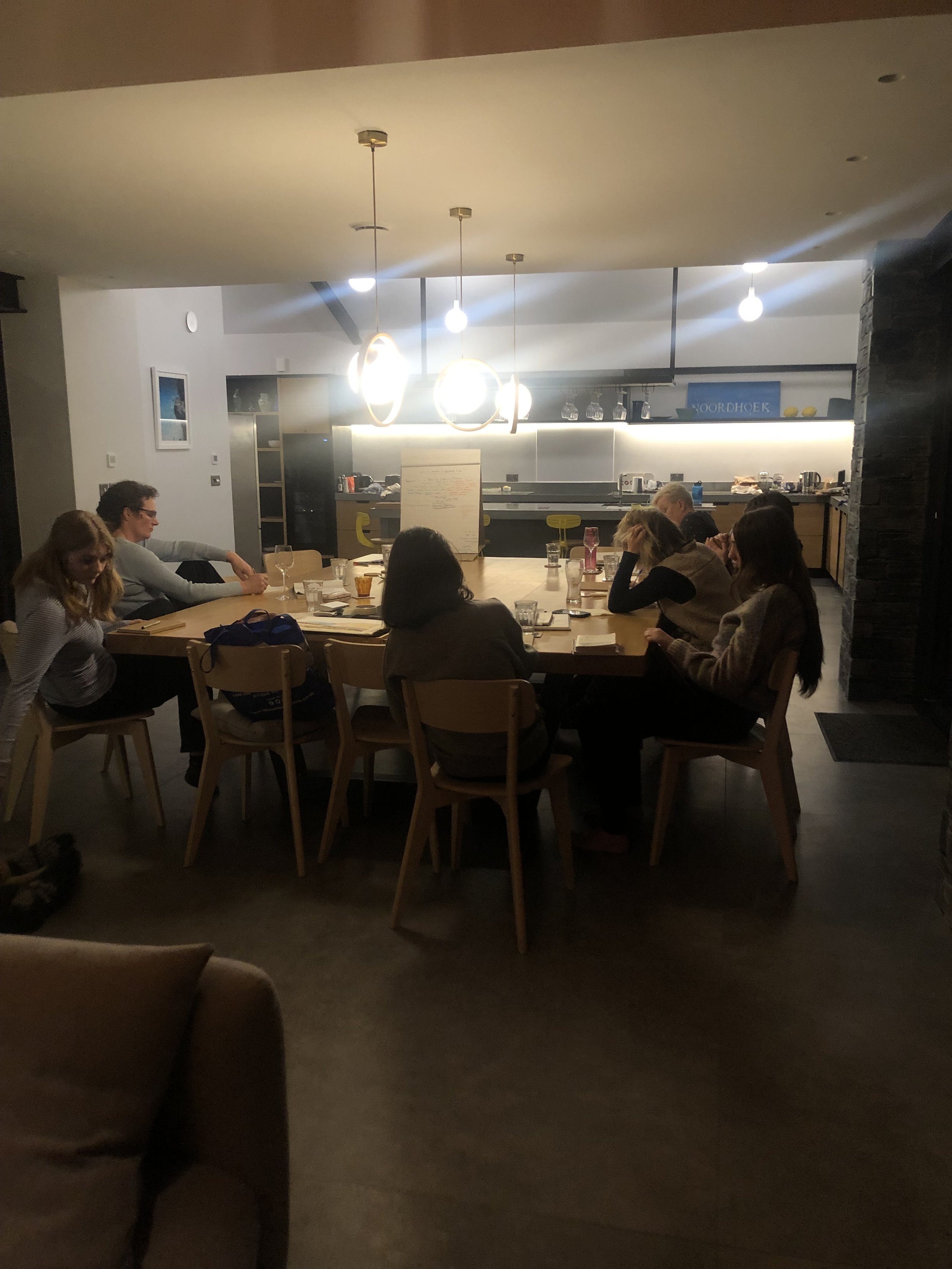 A group of people sitting around a dining table in a modern kitchen, engaging in a discussion or meeting. The room is softly lit with overhead pendant lights, and a whiteboard is placed in the center of the table. The kitchen in the background is sle