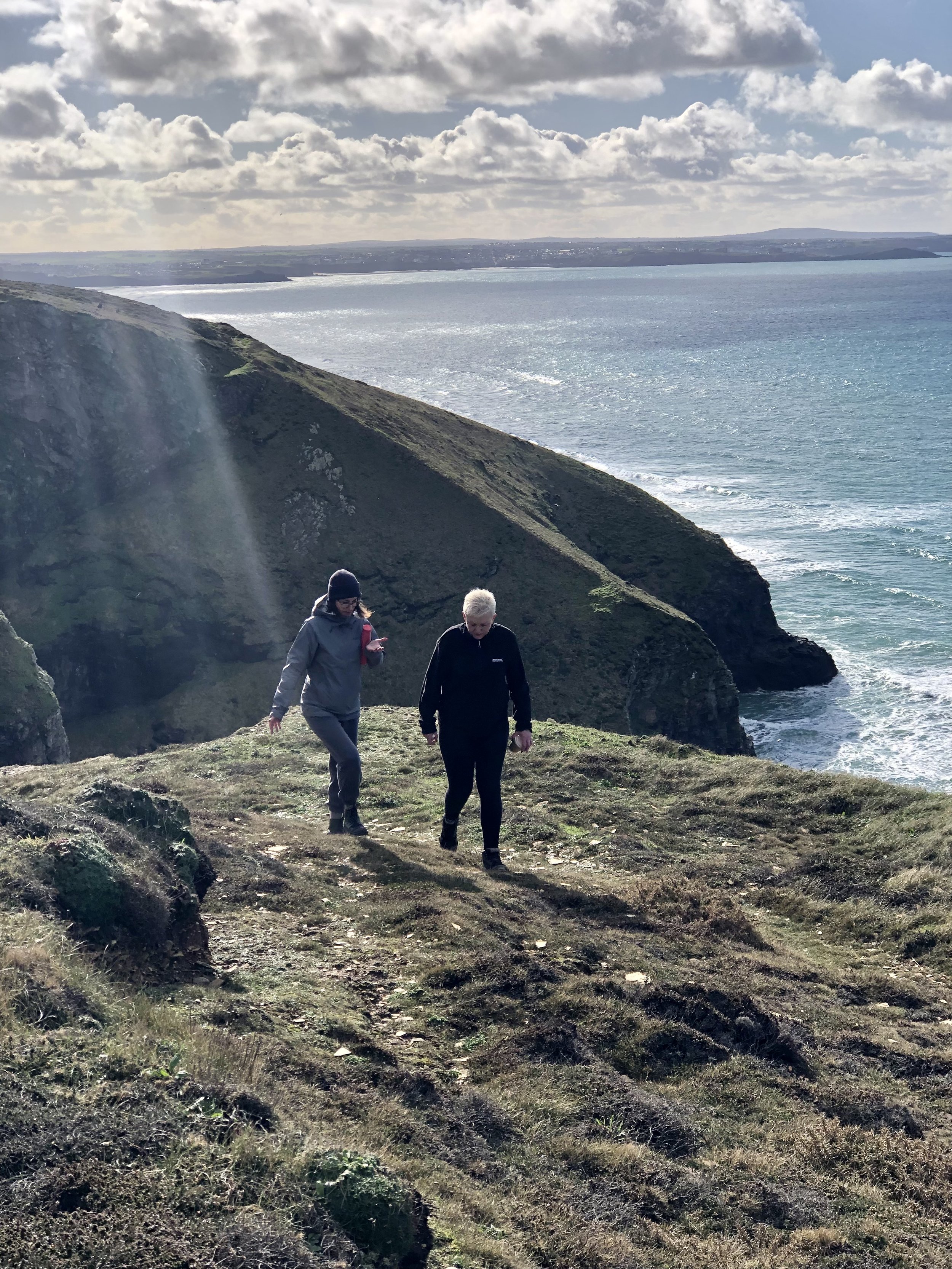 Two people hiking on a grassy cliff overlooking the ocean with clouds in the sky.