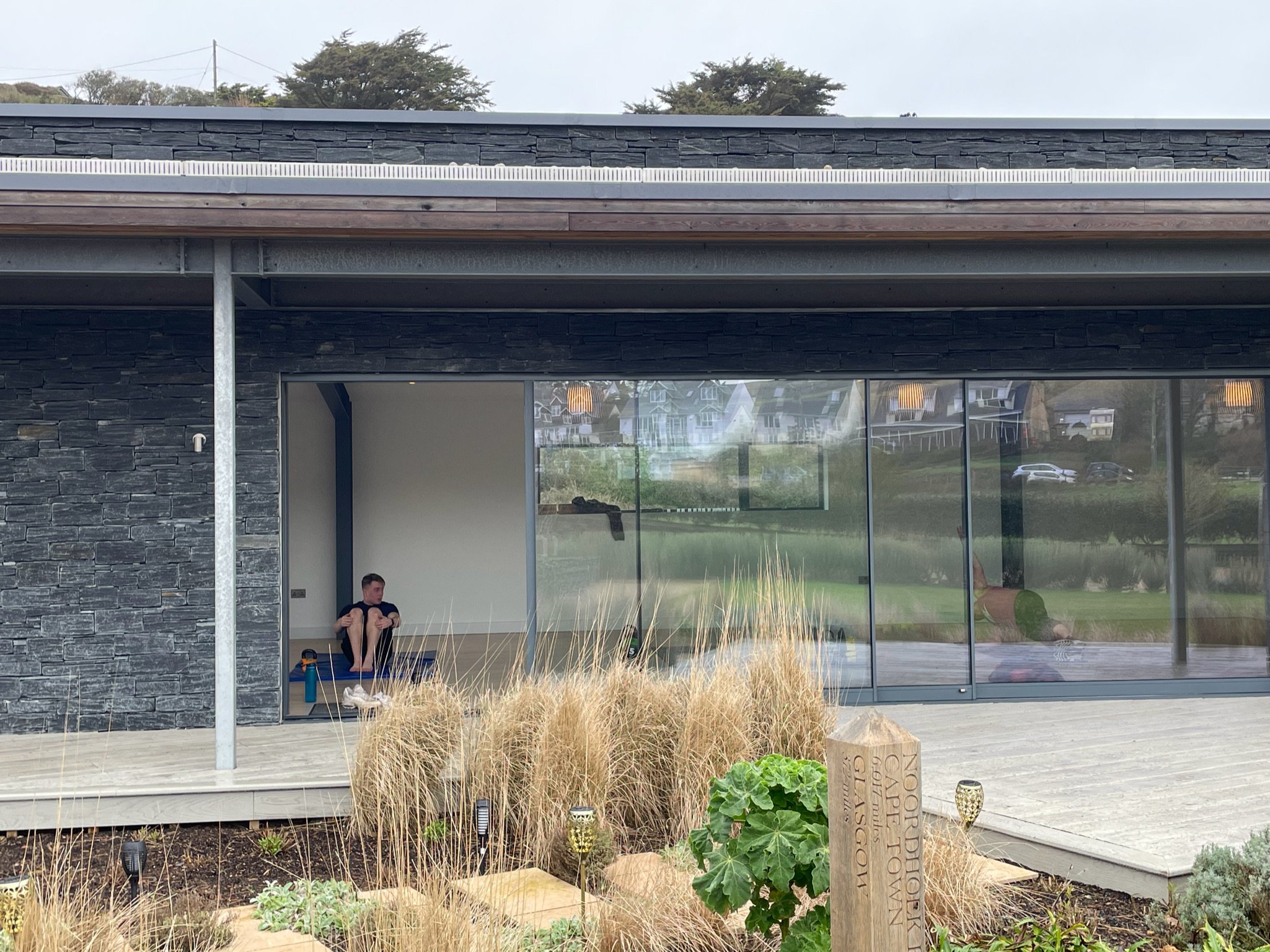 Modern house exterior with stone walls and glass doors, a person sitting in a minimalistic interior, surrounded by landscaped garden with grasses.